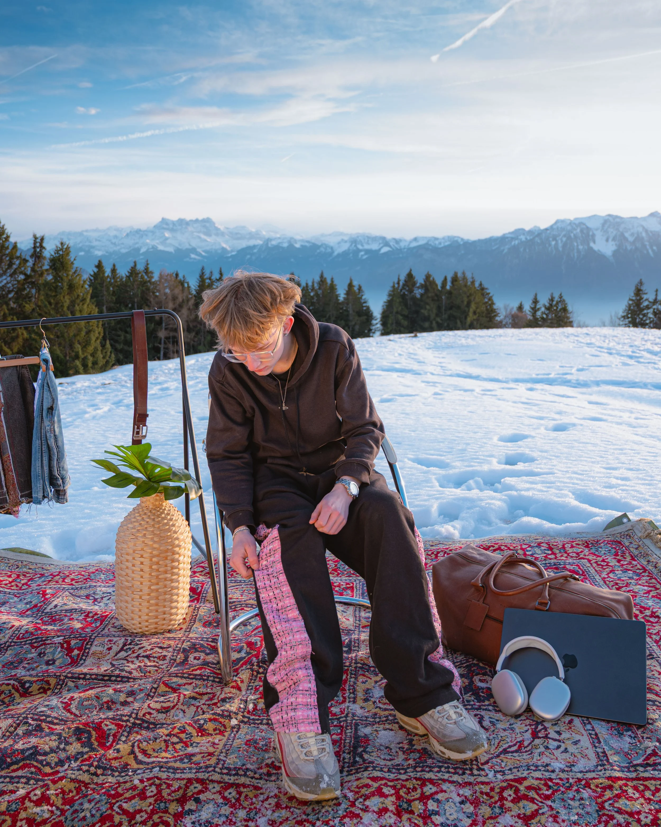 Une personne s'assoit sur un tapis orientale à l'extérieur en montagne enneigée, avec des montagnes et un ciel bleu en arrière-plan. Autour d'elle, il y a des écouteurs, un sac, des vêtements suspendus, et une plante en vase."