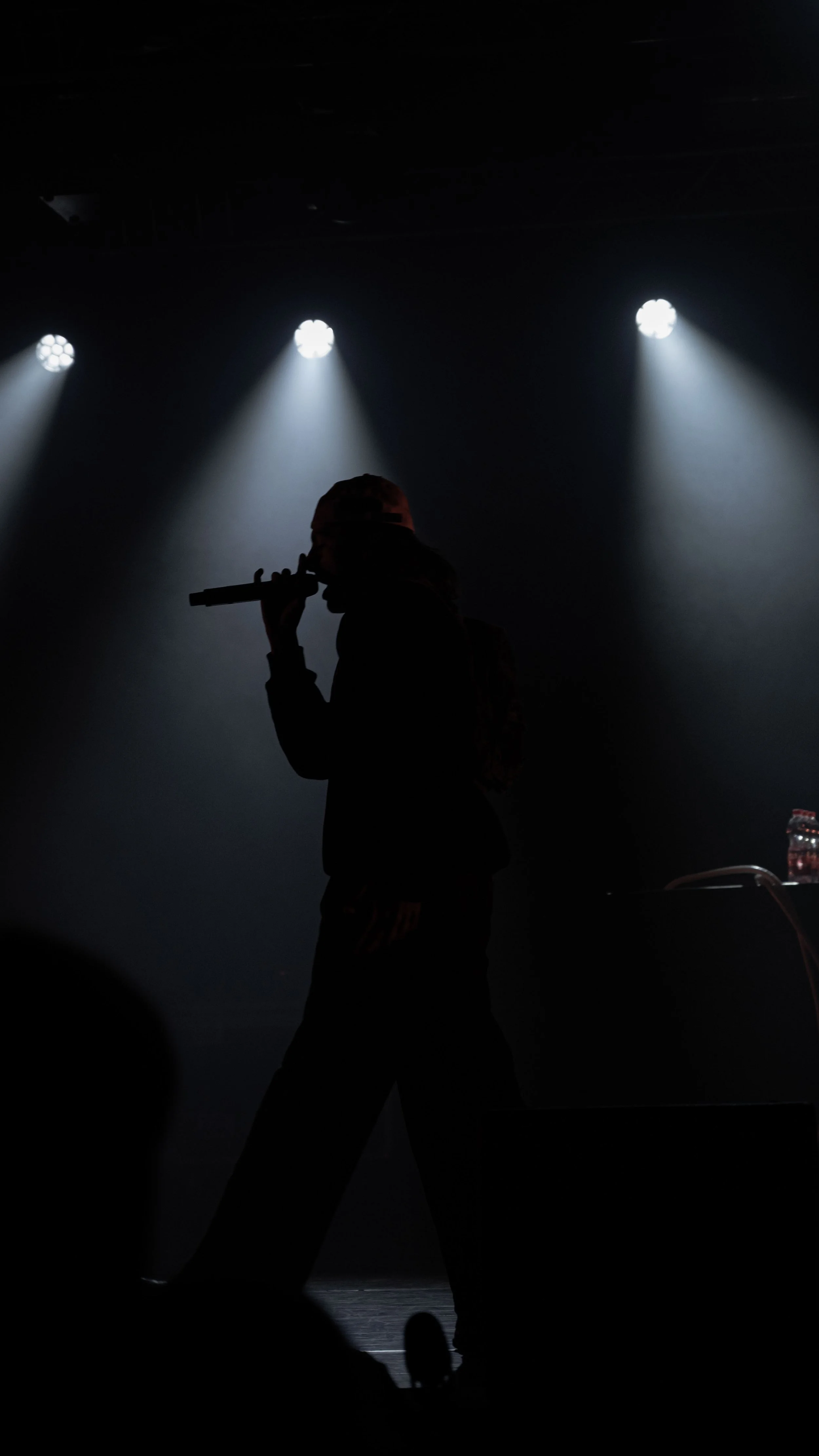 Silhouette d'un enfant avec un chapeau, tenant un microphone sur scène, éclairé par des projecteurs. Style de concert ou de spectacle.