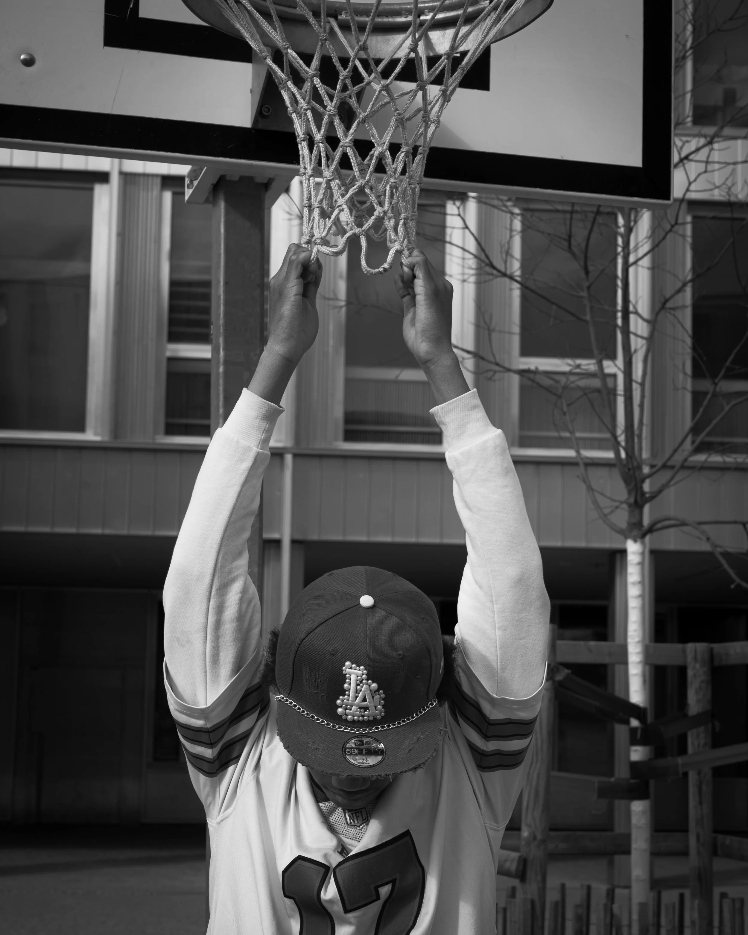 Une personne portant un chapeau de baseball noir avec des perles formant les lettres 'LA' et la numérotation '97', porte un maillot de football américain avec le numéro 92, se tenant devant un panier de basketball sans la balle, en arrière-plan une m