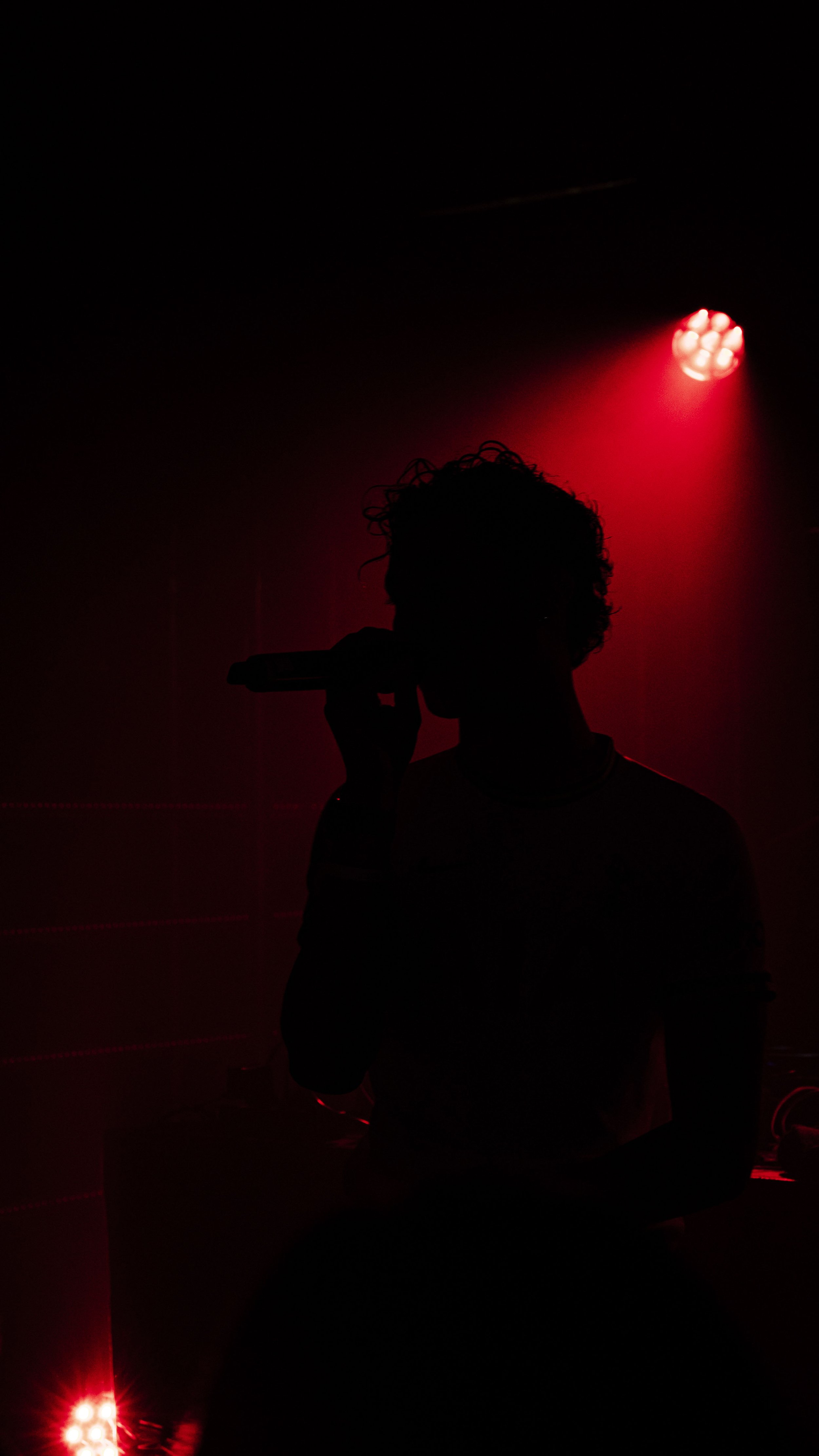 Silhouette d'une personne tenant un microphone sur scène, éclairée par des lumières rouges dans l'obscurité.