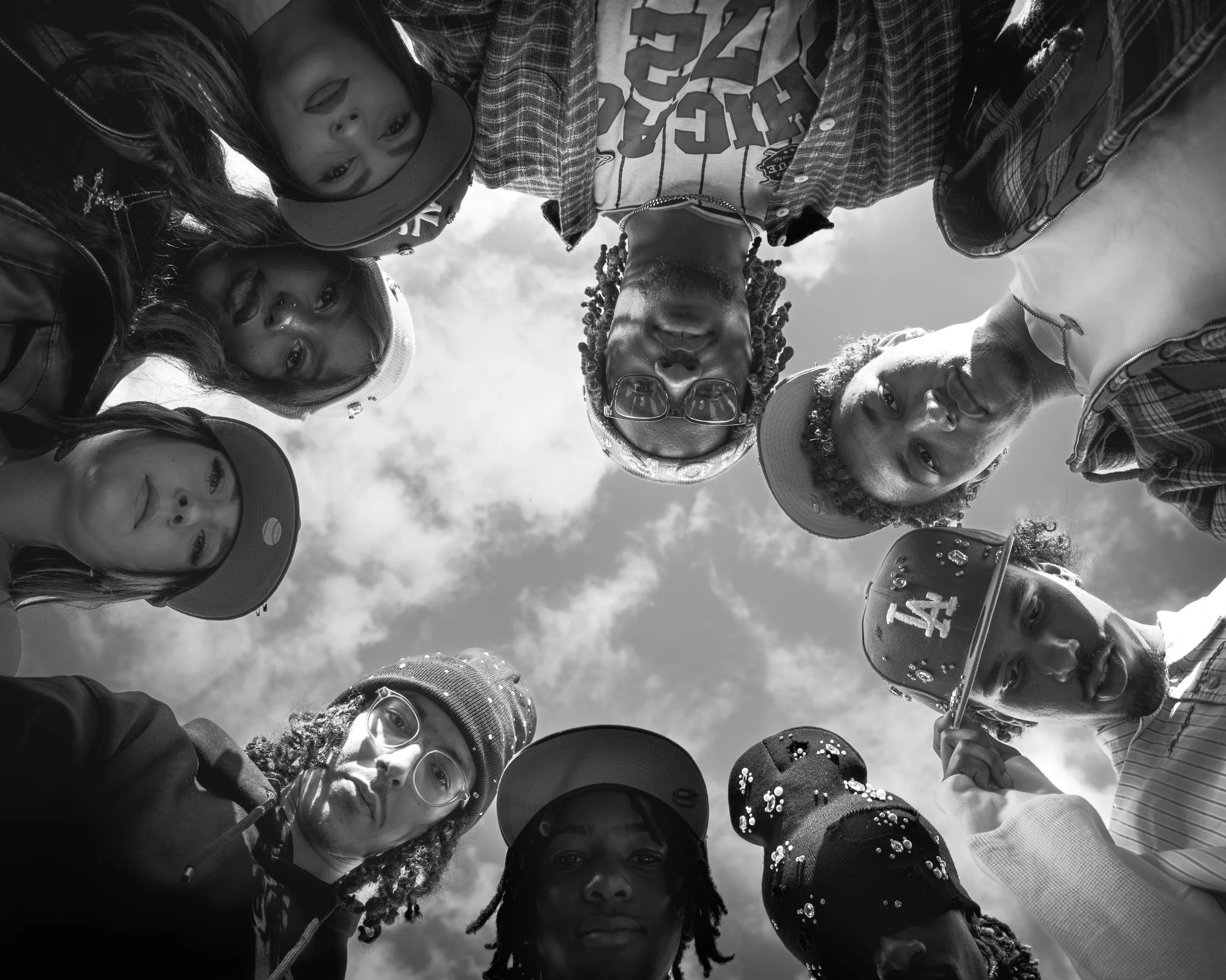 Un groupe diversifié d'enfants et d'adolescents, portant des casquettes et des vêtements décontractés, se regroupent en cercle en regardant vers le bas, sous un ciel nuageux, pris en photo en contre-plongée.