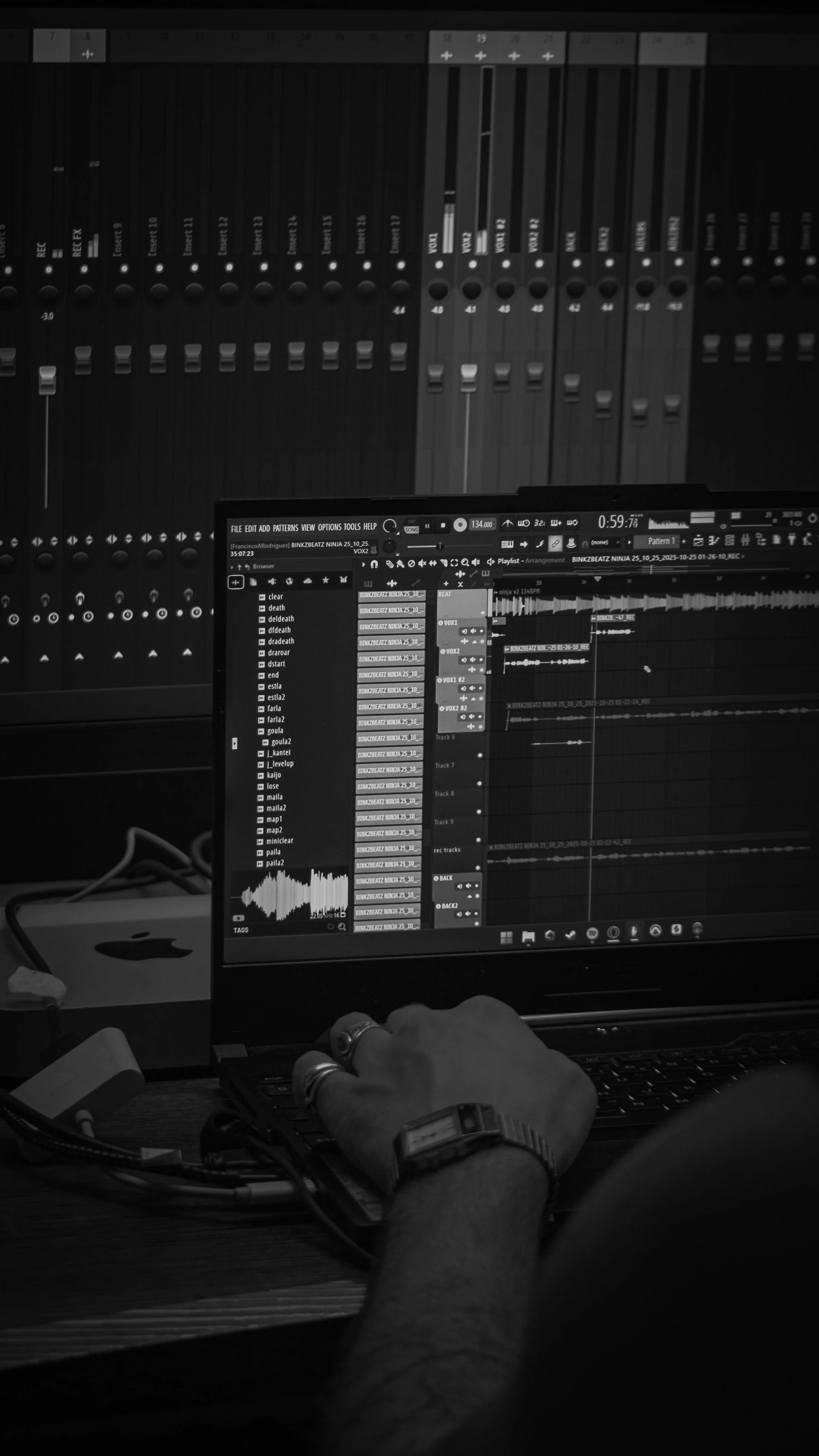 Un poste de travail avec un ordinateur portable affichant un logiciel de production musicale, en face d'une table de mixage audio, avec une main portant une montre et plusieurs bagues au premier plan.