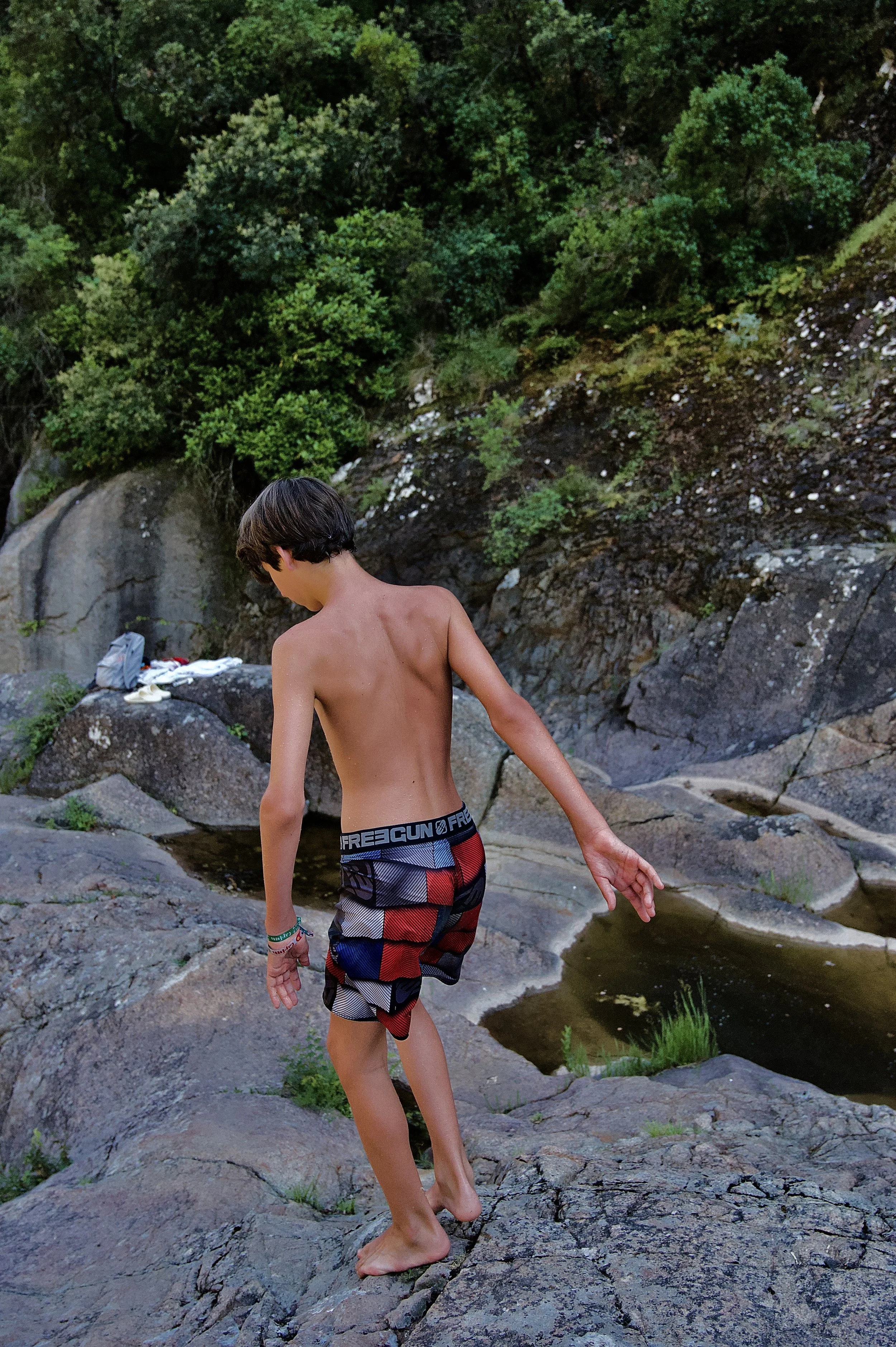 Jeune garçon avec un short à motif, debout sur des rochers près d'un cours d'eau dans une nature verdoyante.