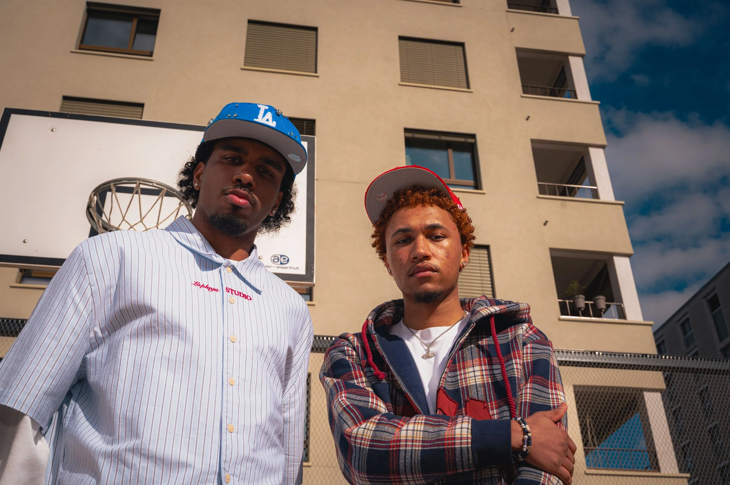 Deux jeunes hommes posent devant un bâtiment résidentiel avec un panneau de basketball en arrière-plan, portant des casquettes et des vêtements décontractés.
