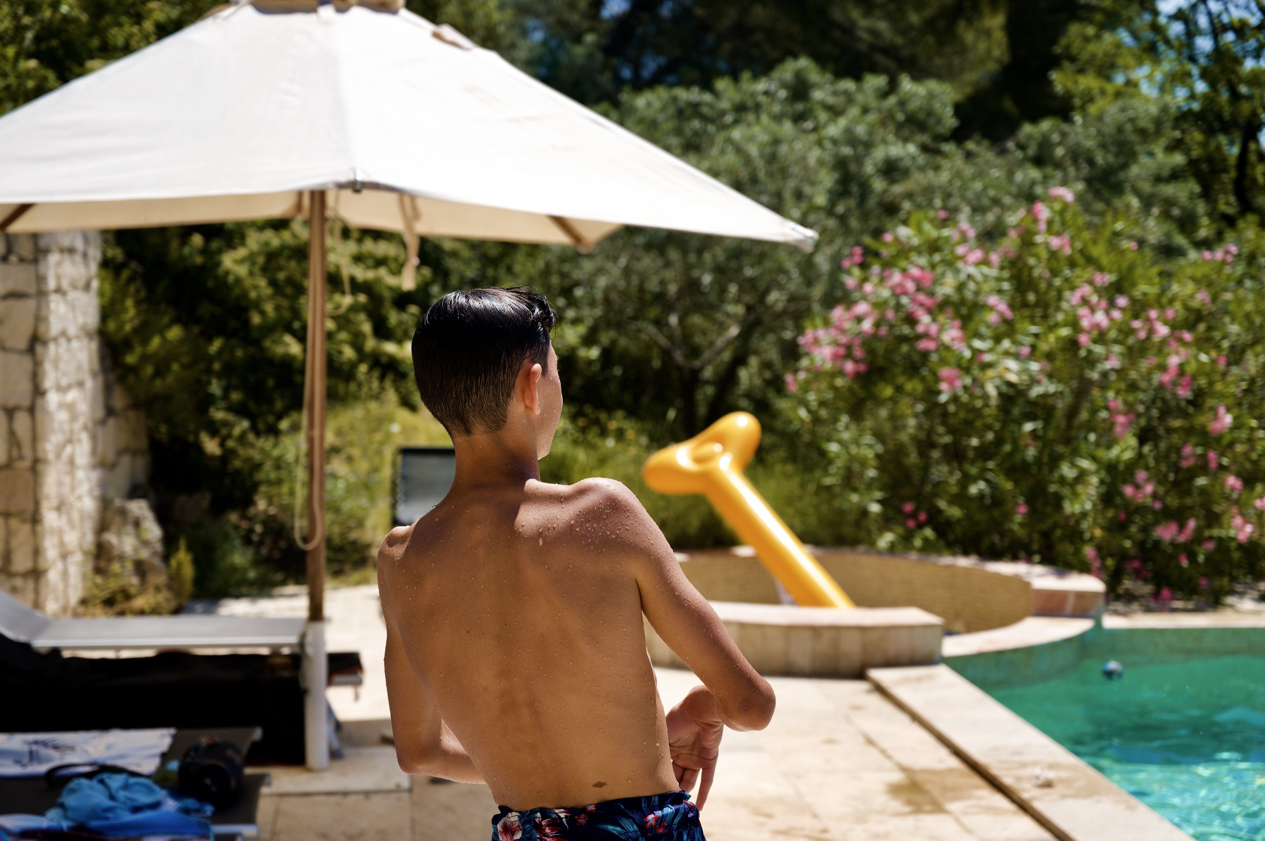 Un homme nu vu de dos, près d'une piscine, avec un parasol blanc en arrière-plan, entouré de végétation verte et de fleurs roses.