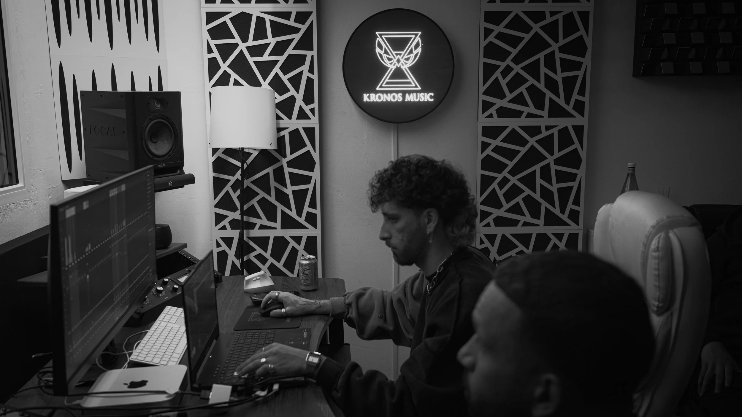 Une personne avec des cheveux bouclés et un tatouage sur le bras, assise devant un ordinateur dans un studio d'enregistrement ou de production musicale, avec un mur décoratif en arrière-plan et un logo lumineux 'KRONOS MUSIC'.
