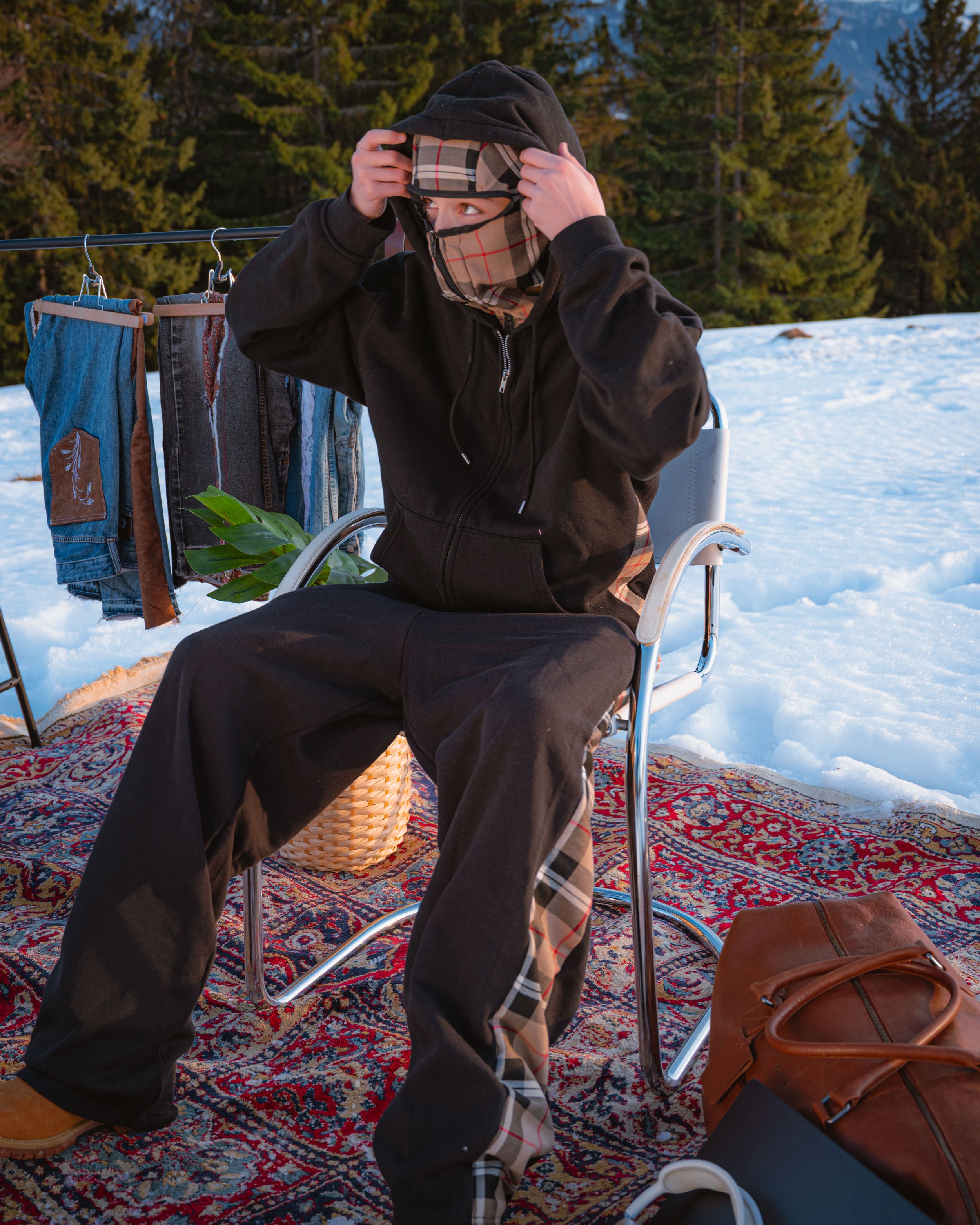 Une personne dans un fauteuil pliant, portant un cache-visage à motif tartan, ajuste une capuche sur la tête, dans un environnement enneigé entouré d'arbres forestiers.