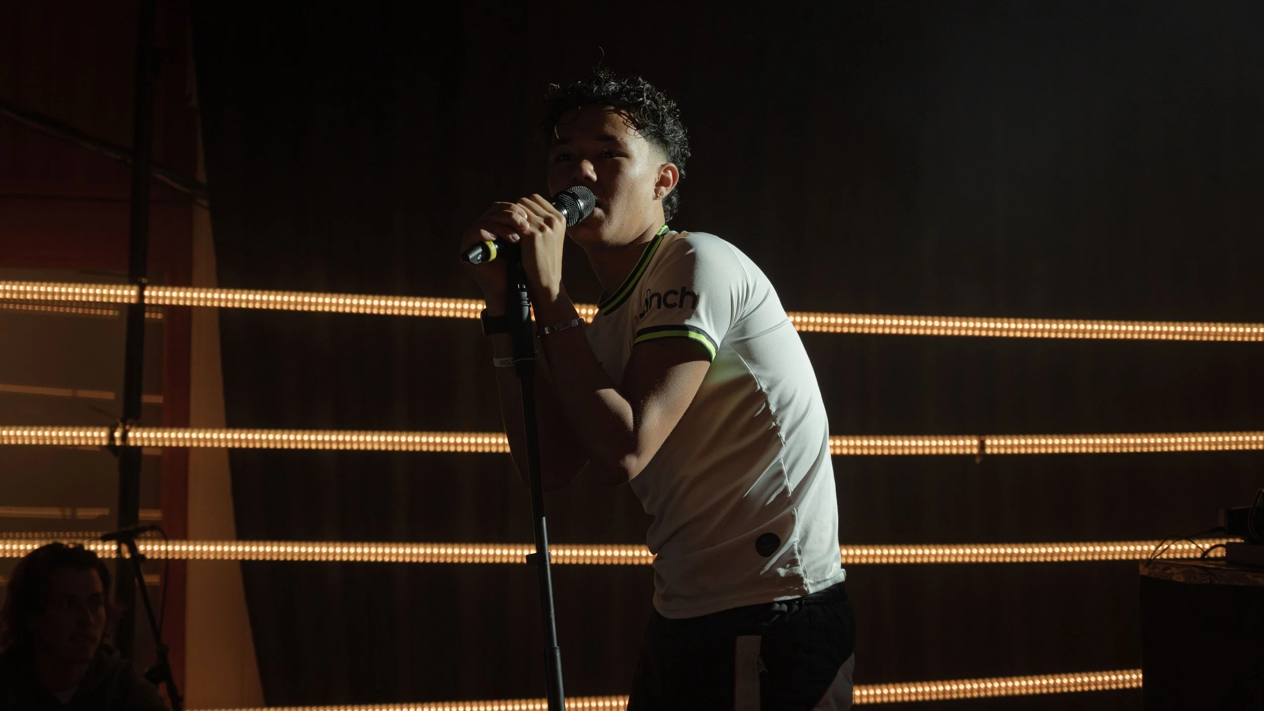 Un jeune homme chante dans un micro, éclairé par des lumières en arrière-plan.
