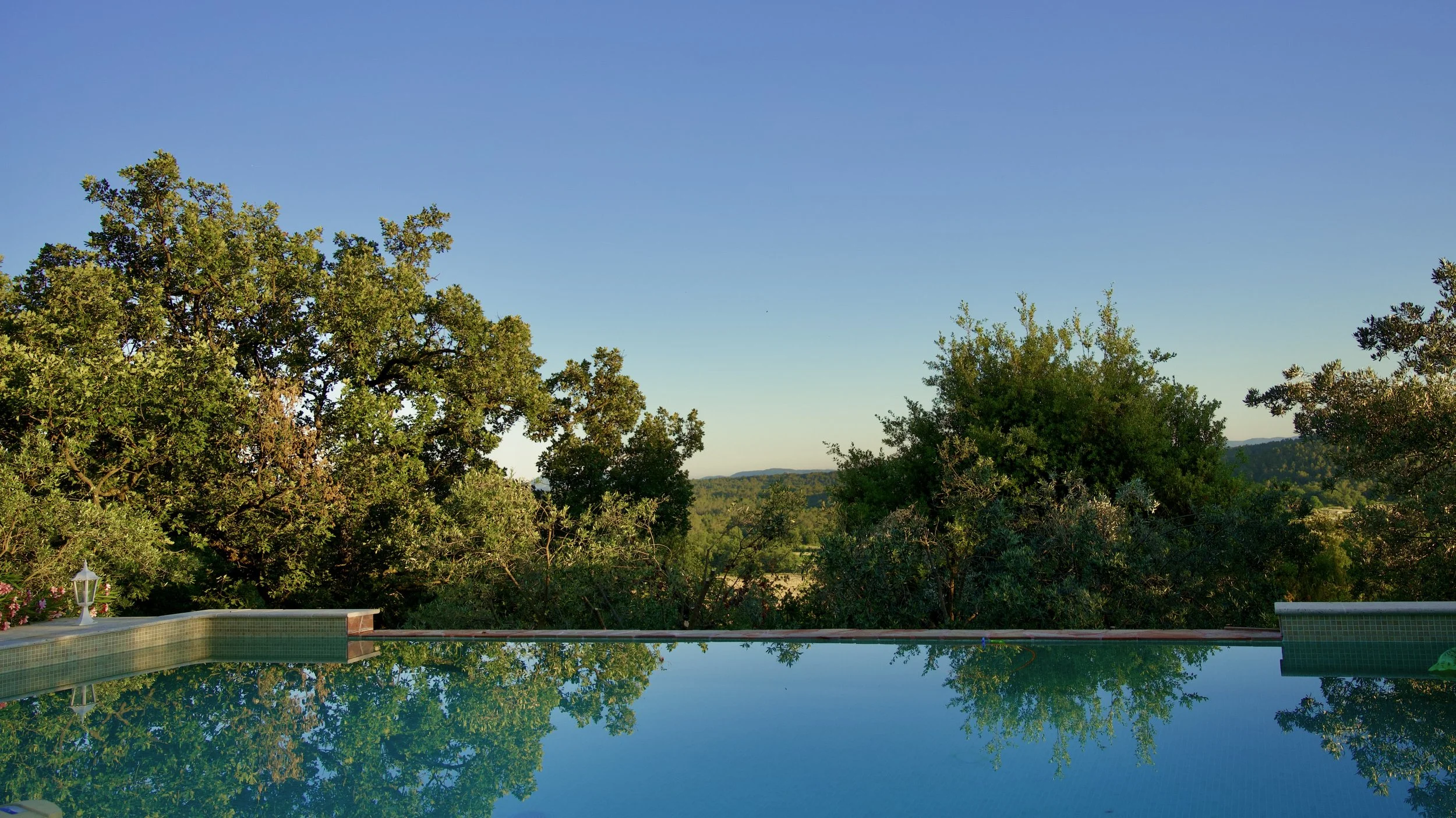 Une piscine reflète le ciel bleu et les arbres verts entourant la zone, avec des collines au loin.