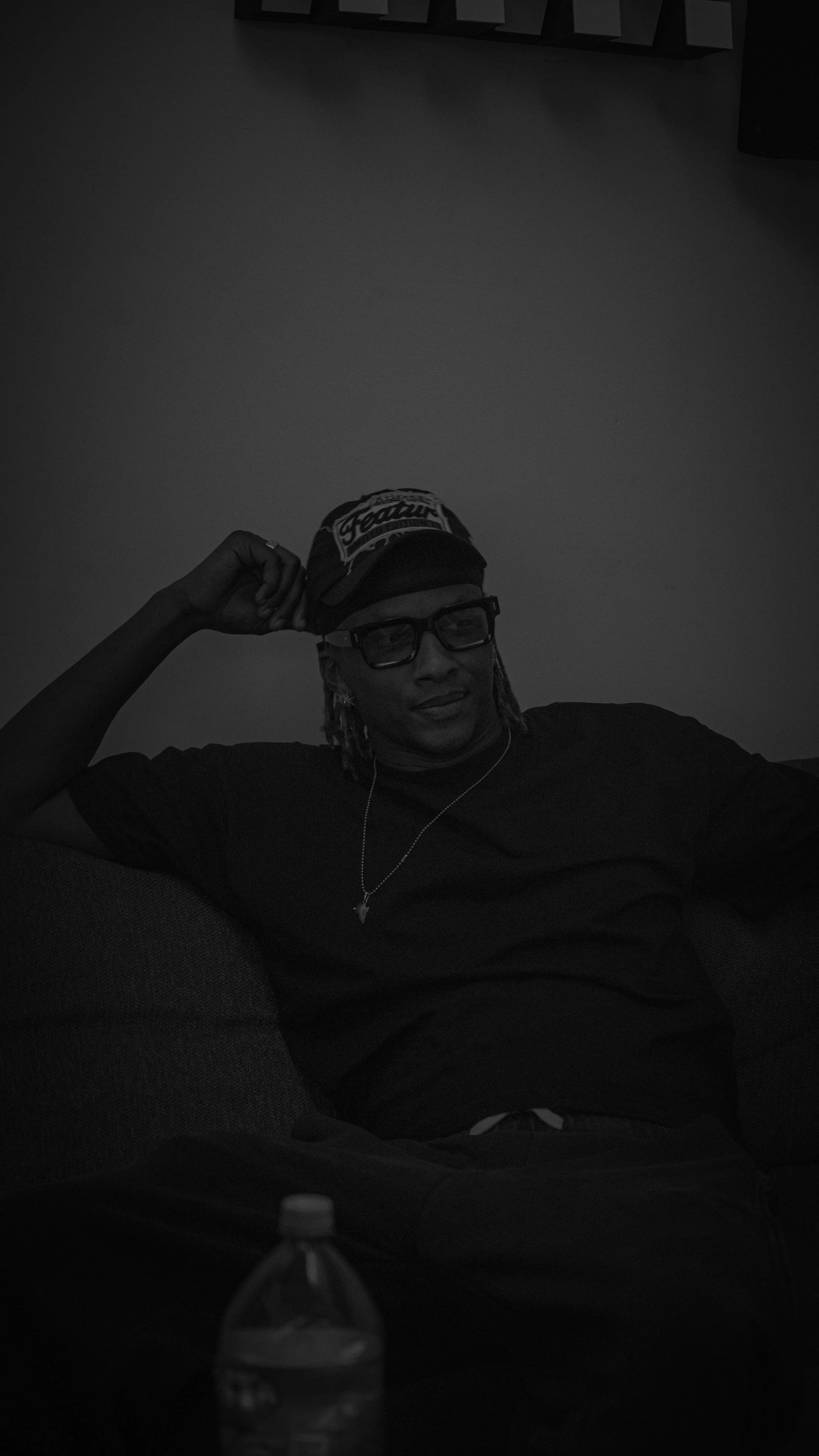 Un homme assis sur un canapé portant un t-shirt noir, des lunettes noires et un bonnet. Il sourit légèrement et tient un panneau ou un vêtement sur sa tête. Un bouteille d'eau est visible à l'avant-plan.