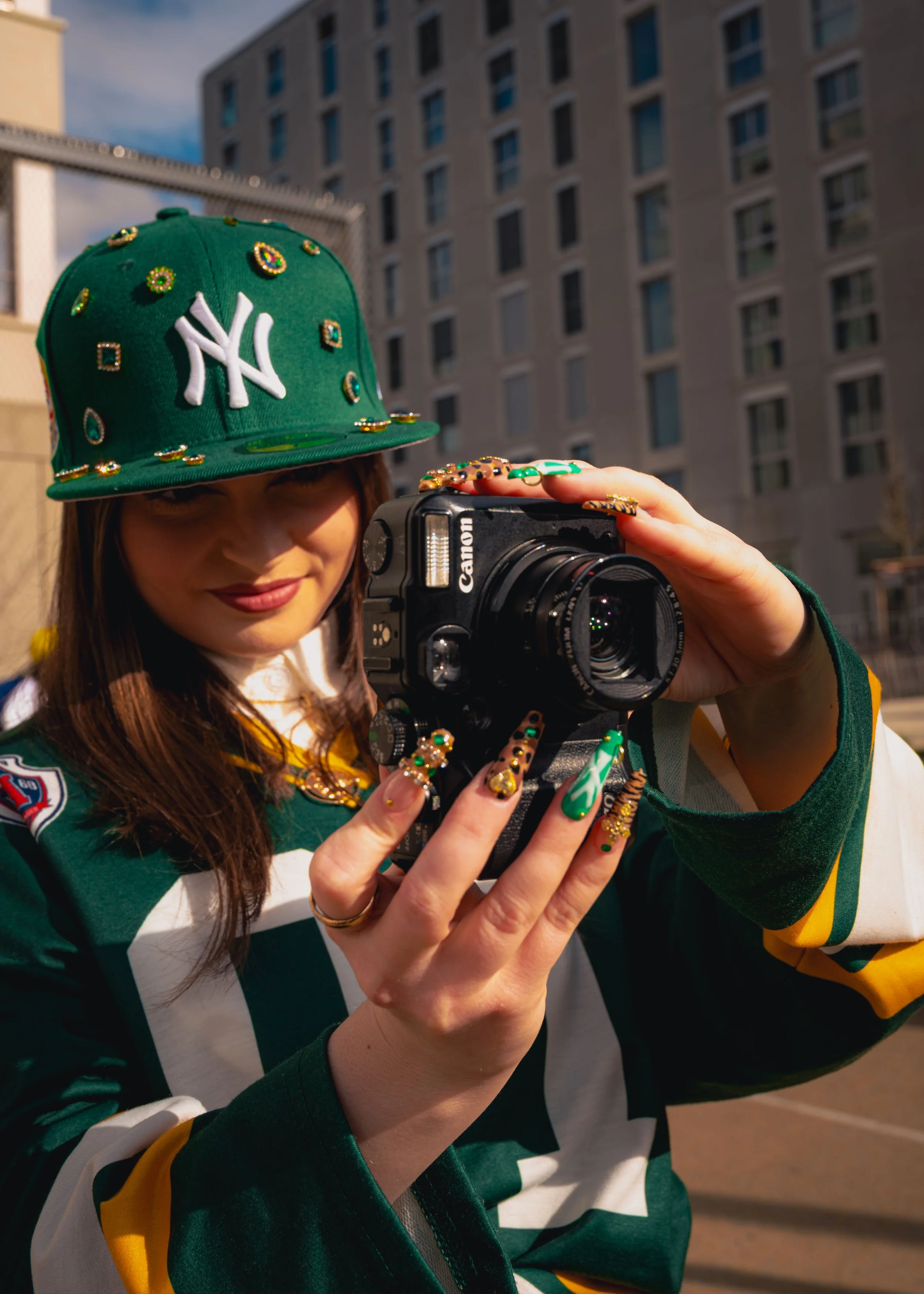 Jeune femme portant une casquette verte décorée de bijoux, prenant un selfie avec un appareil photo, en plein air dans une ville.