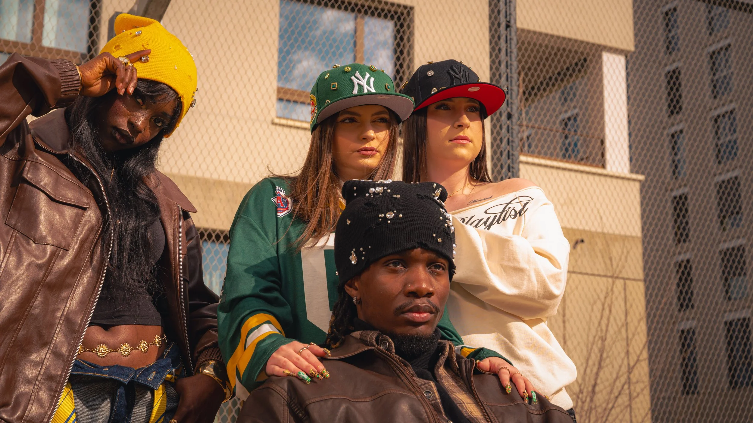 Groupe de jeunes adultes portant des casquettes, debout devant un mur avec une clôture, dans un environnement urbain.