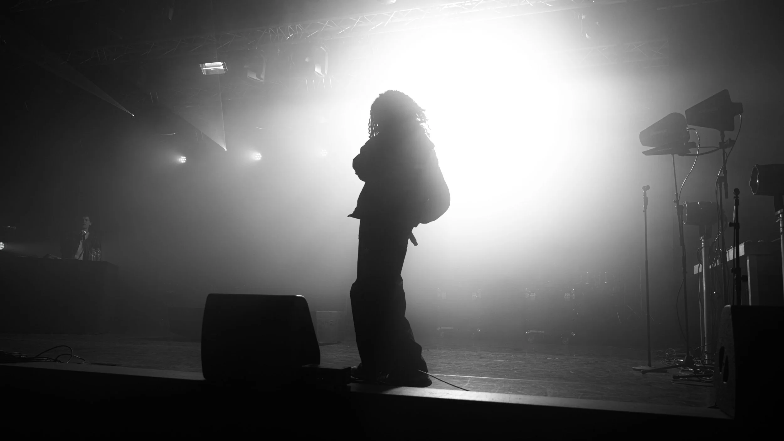 Silhouette d'une personne debout sur scène avec forte lumière en arrière-plan, équipements de scène visibles sur le côté droit.