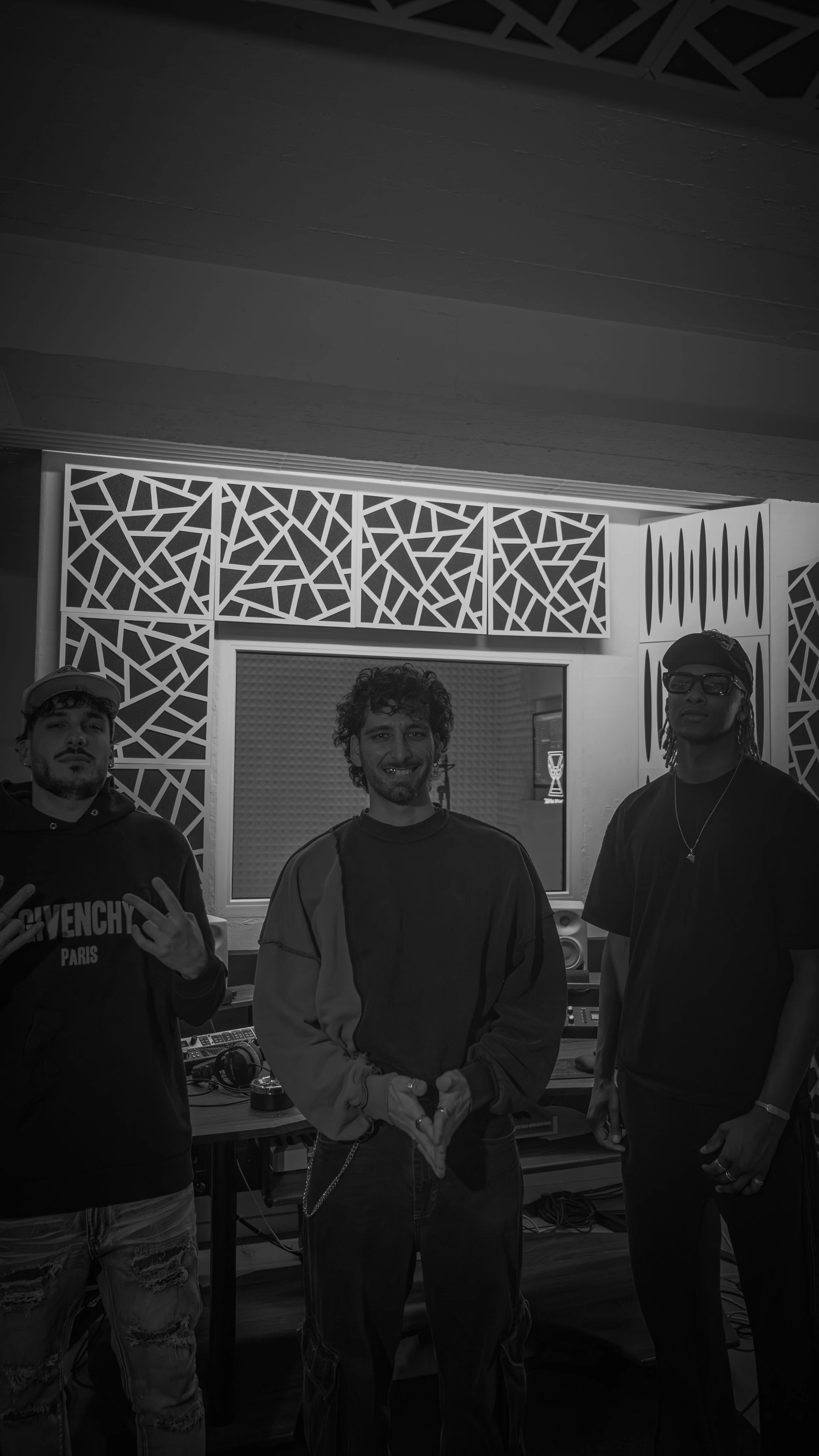 Trois hommes posent dans un studio d'enregistrement ou de musique. Un homme au centre fait un geste de main en forme de cœur, les autres hommes portent des vêtements décontractés.