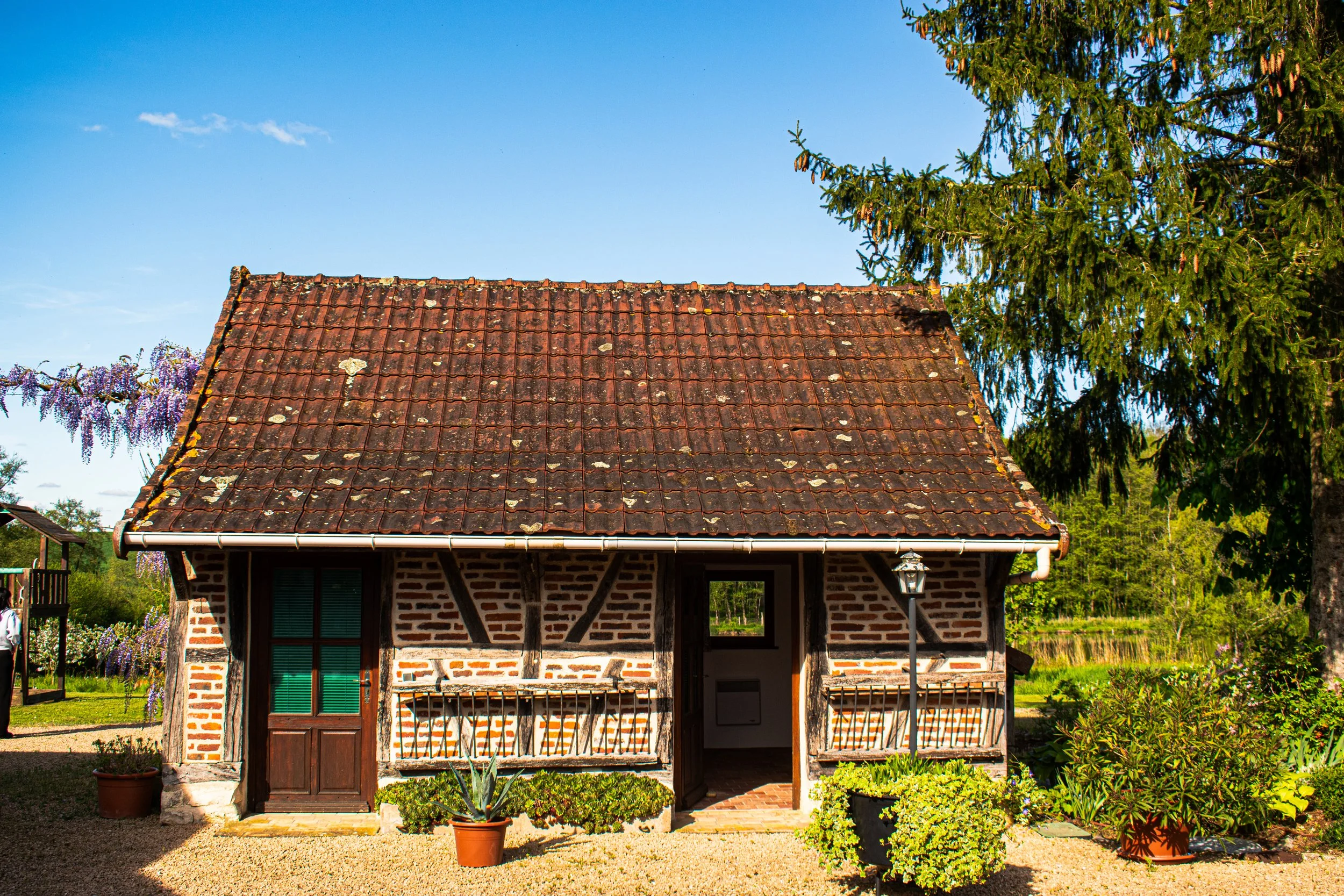 Maison en briques avec toiture en tuiles rouges, devant un jardin avec des pots de plantes, sous un ciel bleu clair.