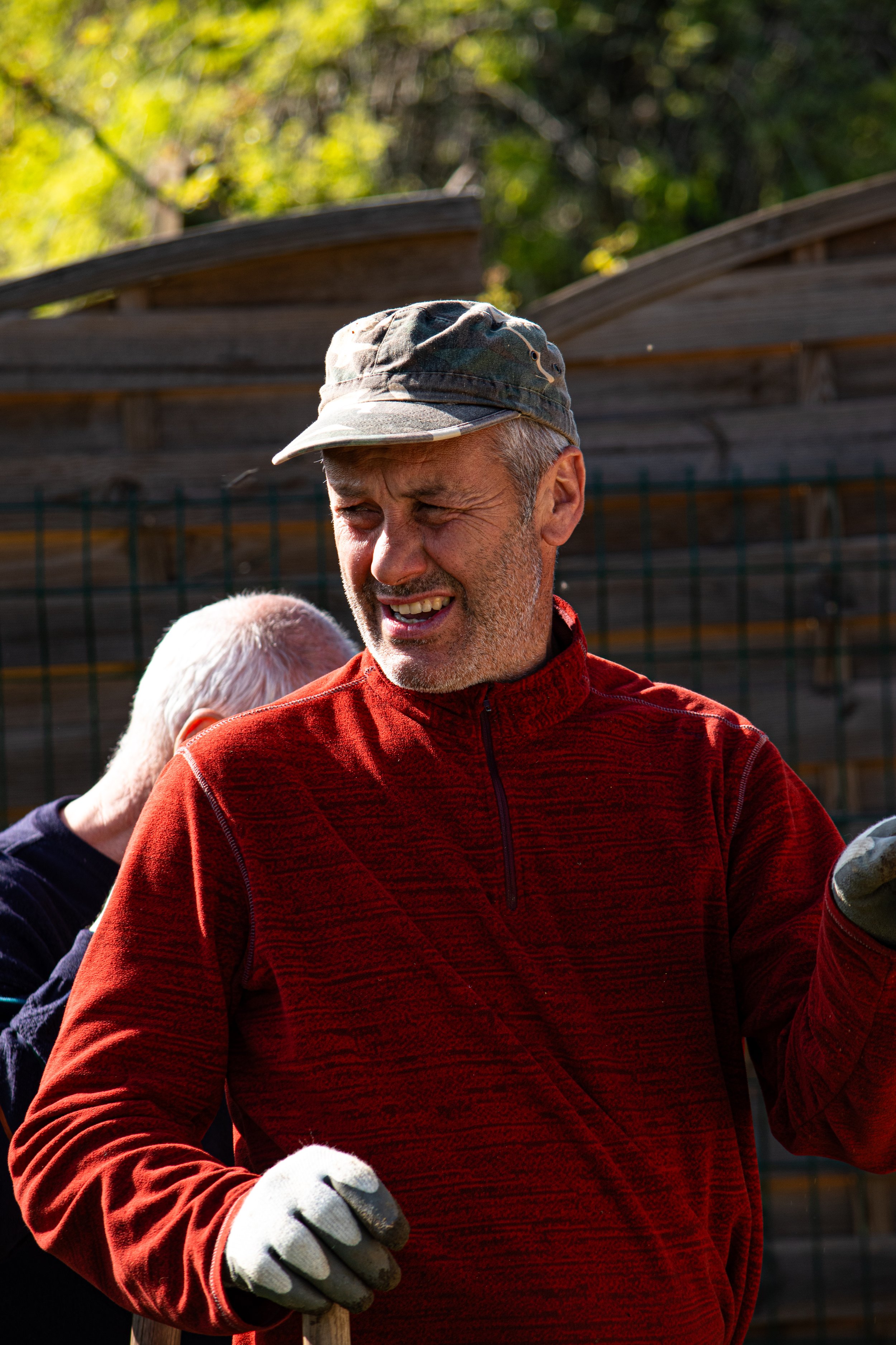 Un homme portant une casquette et un manteau rouge, souriant, en plein air, à côté d'une clôture en métal et d'une structure en bois, sous un arrière-plan de verdure.