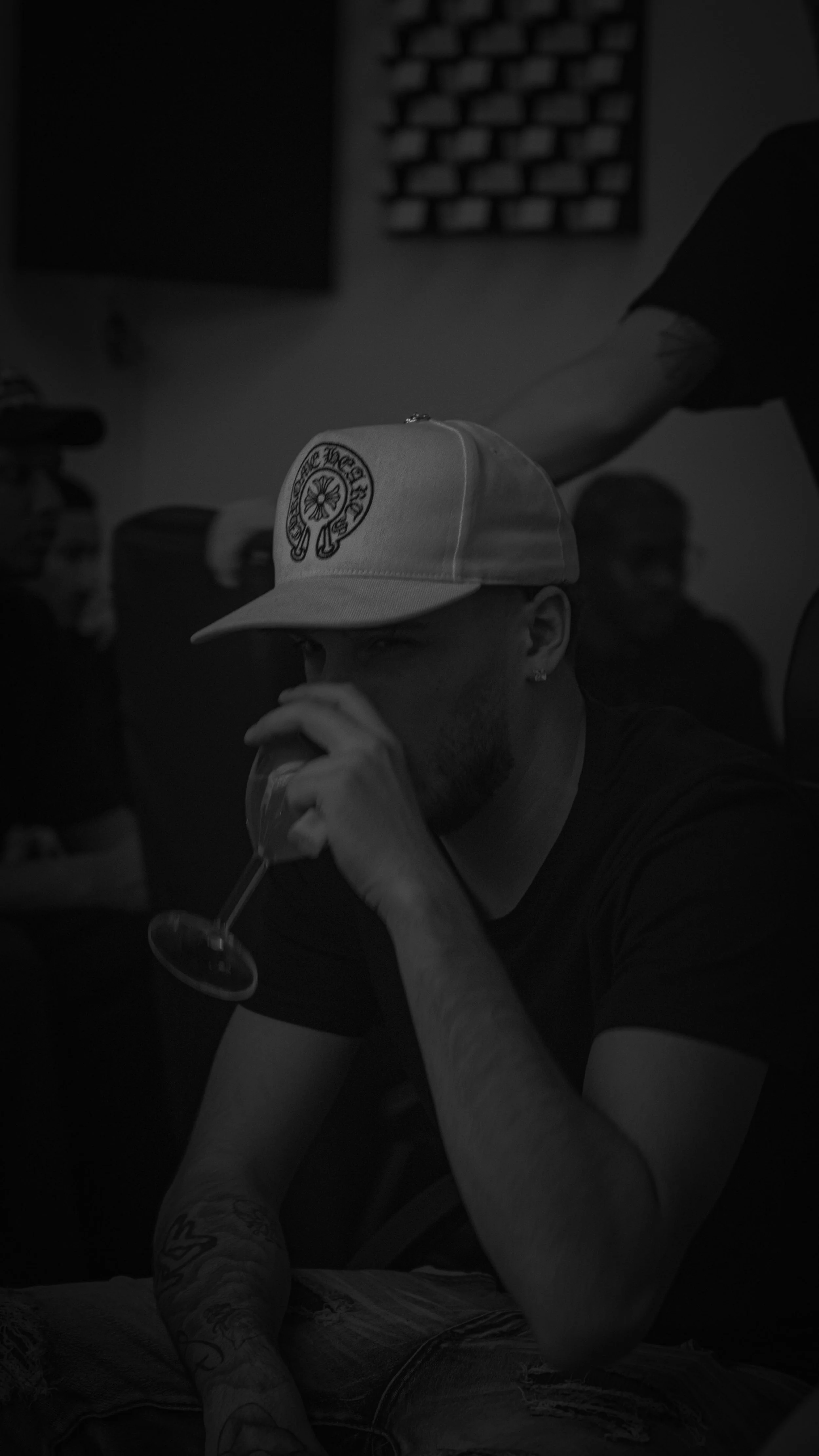 Un homme portant une casquette blanche avec un logo, assis à une table, buvant dans un verre à vin, dans un environnement sombre.