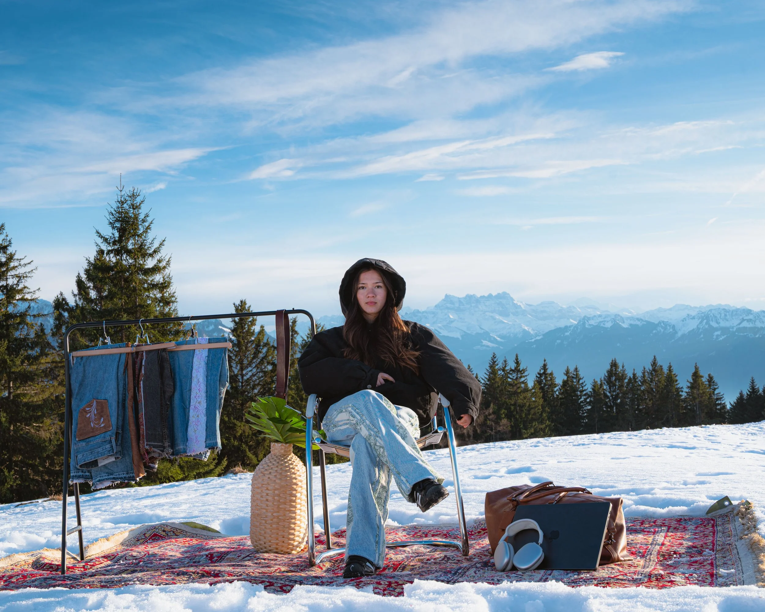 Une jeune femme assise sur une chaise en plein air dans un paysage de montagnes enneigées, portant une veste noire et un chapeau à capuche, entourée d'objets de camping, avec un étendoir à vêtements, des sacs, une tappisserie et un panier en rotin, s
