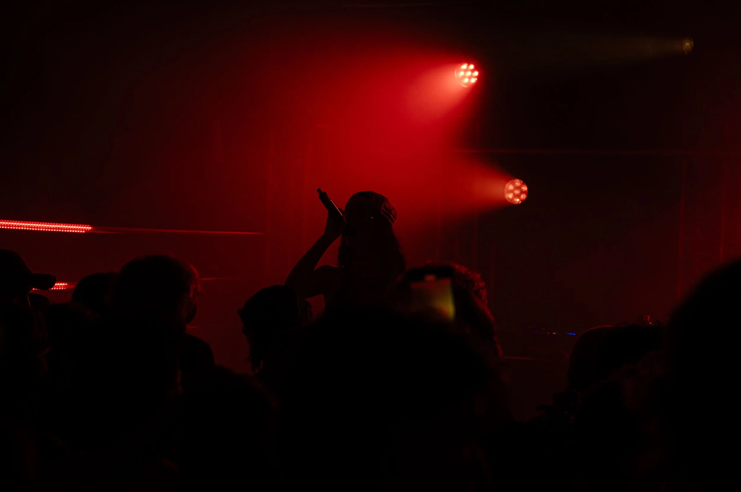 Une silhouette d'une personne en train de prendre une photo ou de regarder vers une scène avec des lumières rouges dans une pièce sombre, avec plusieurs autres silhouettes présentes.