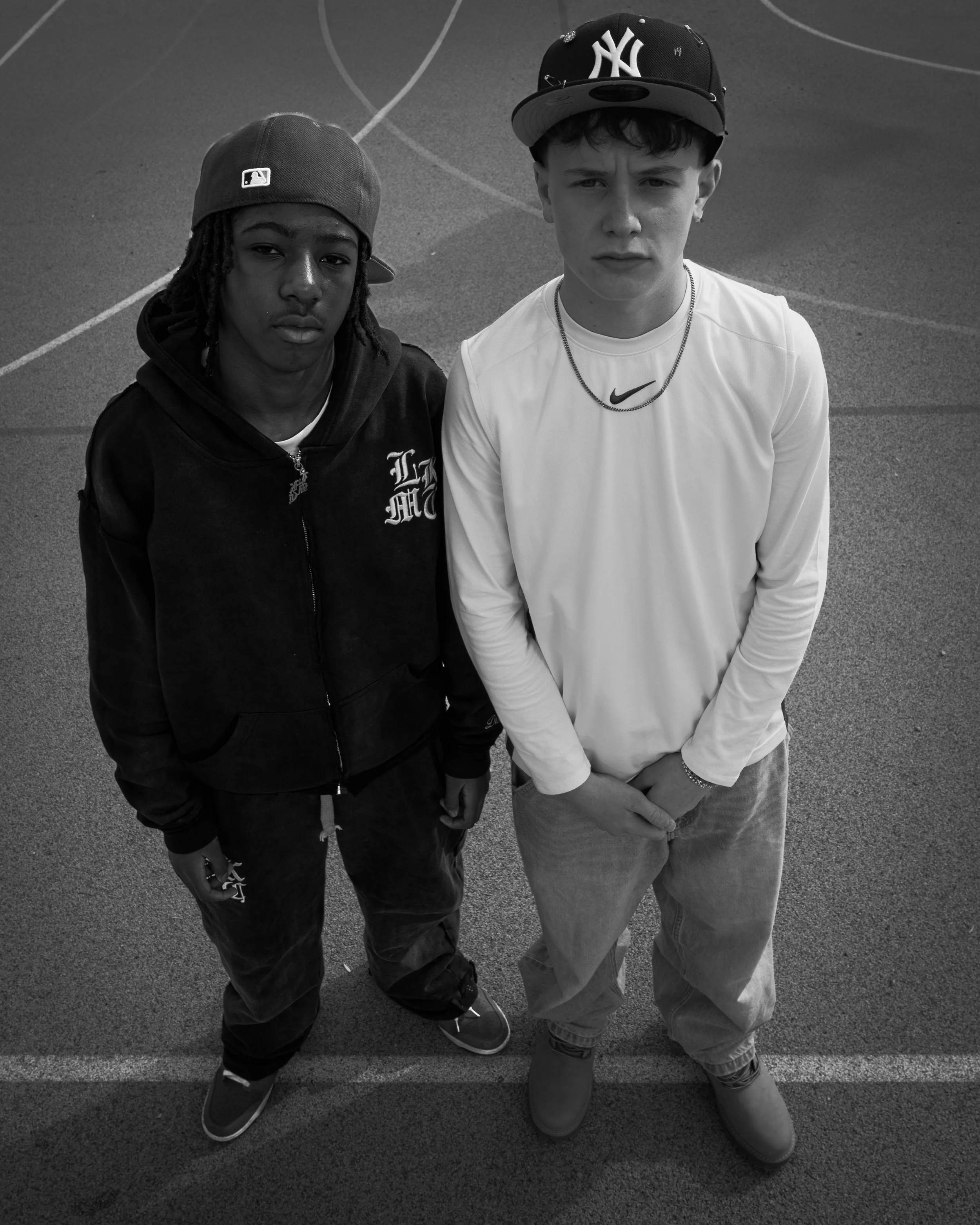 Deux jeunes hommes posent côte à côte sur une surface extérieure, portant des casquettes et des vêtements décontractés.