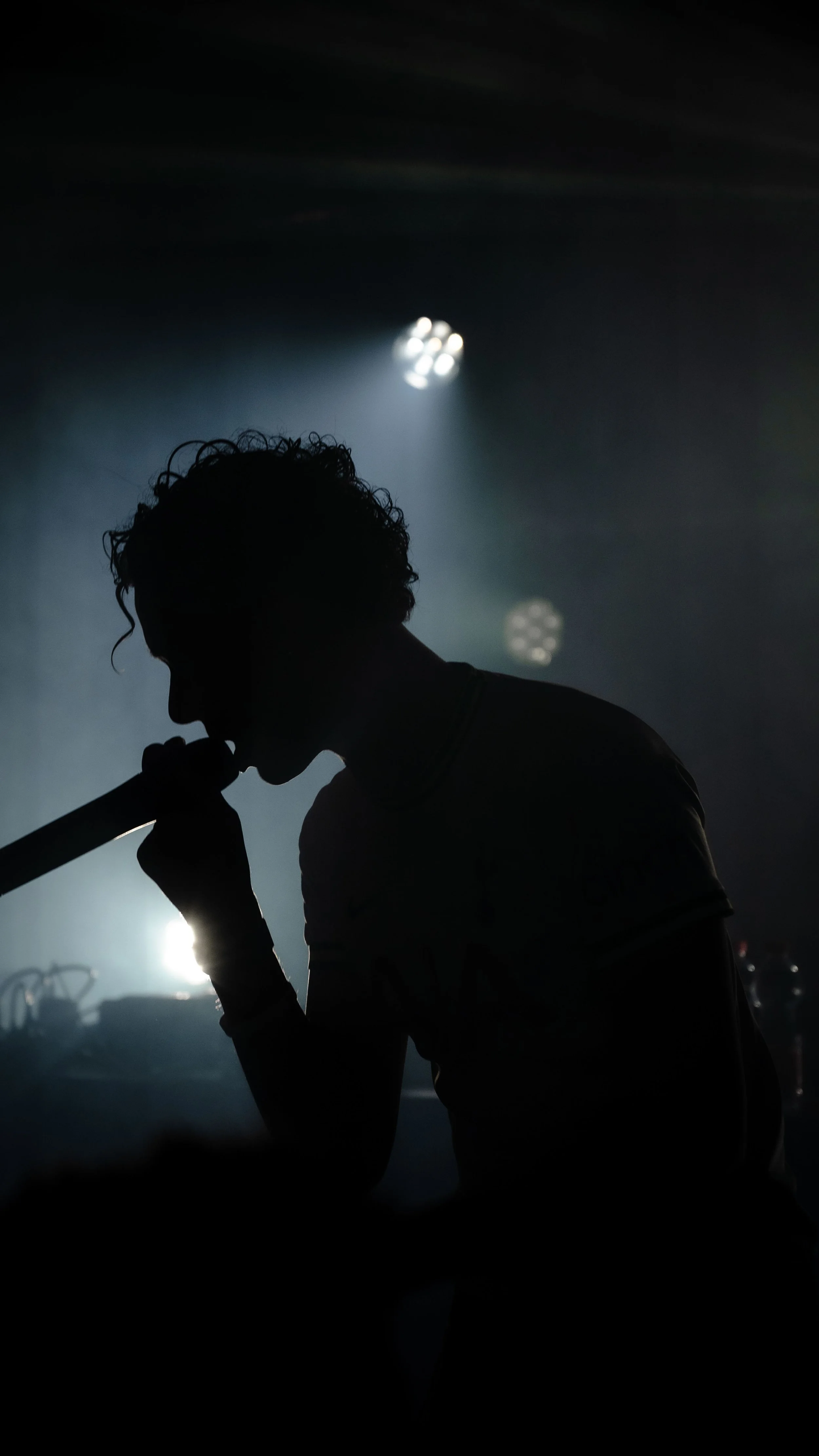 Silhouette d'une personne avec une coiffure bouclée, jouant d'un instrument à vent sur scène, éclairée par des lumières fortes et une ambiance sombre.