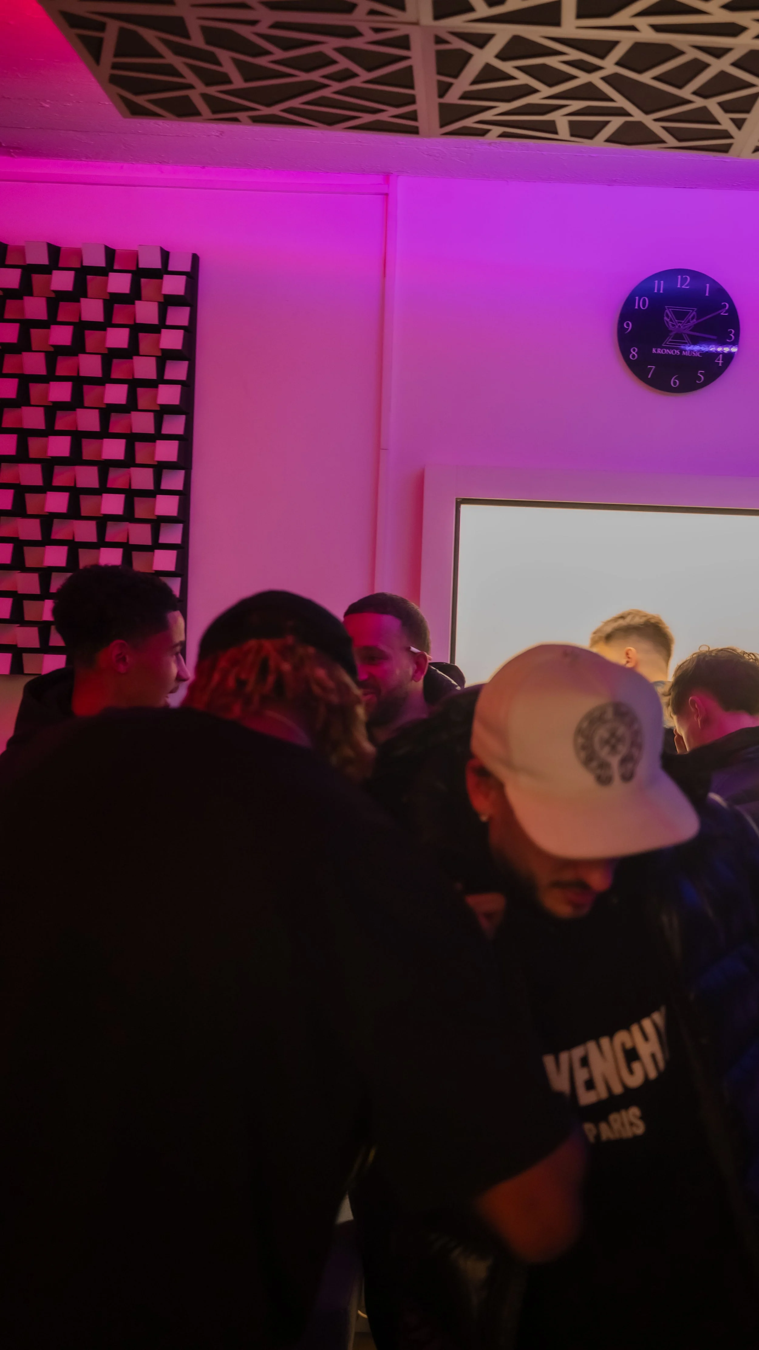 Groupe de personnes dans une pièce avec un éclairage violet et un mur decoratif, certaines portant des casquettes, dans une ambiance sociale ou festive.
