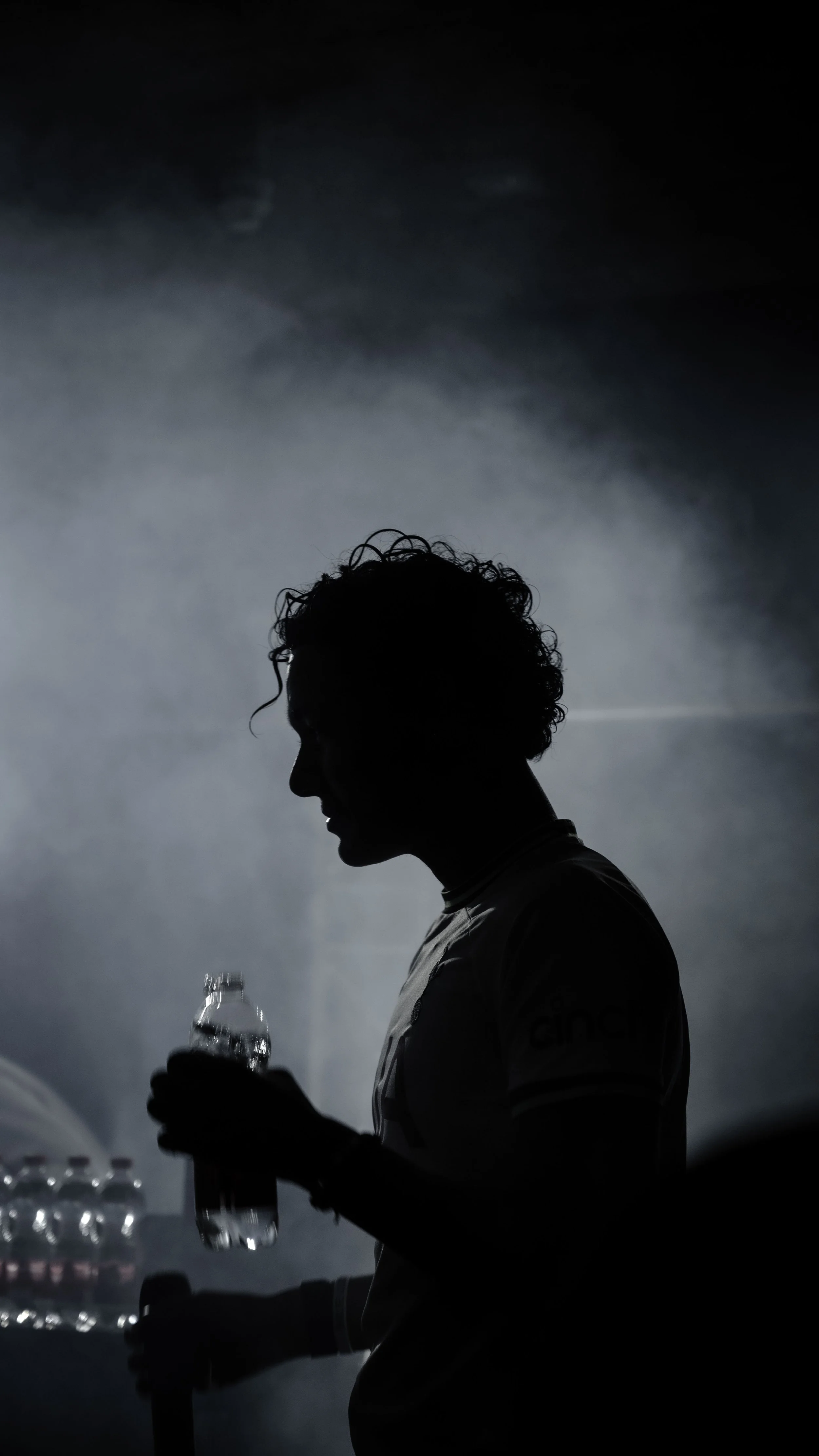 Silhouette d'une personne avec des cheveux bouclés, tenant une bouteille d'eau, dans un environnement sombre et brumeux.