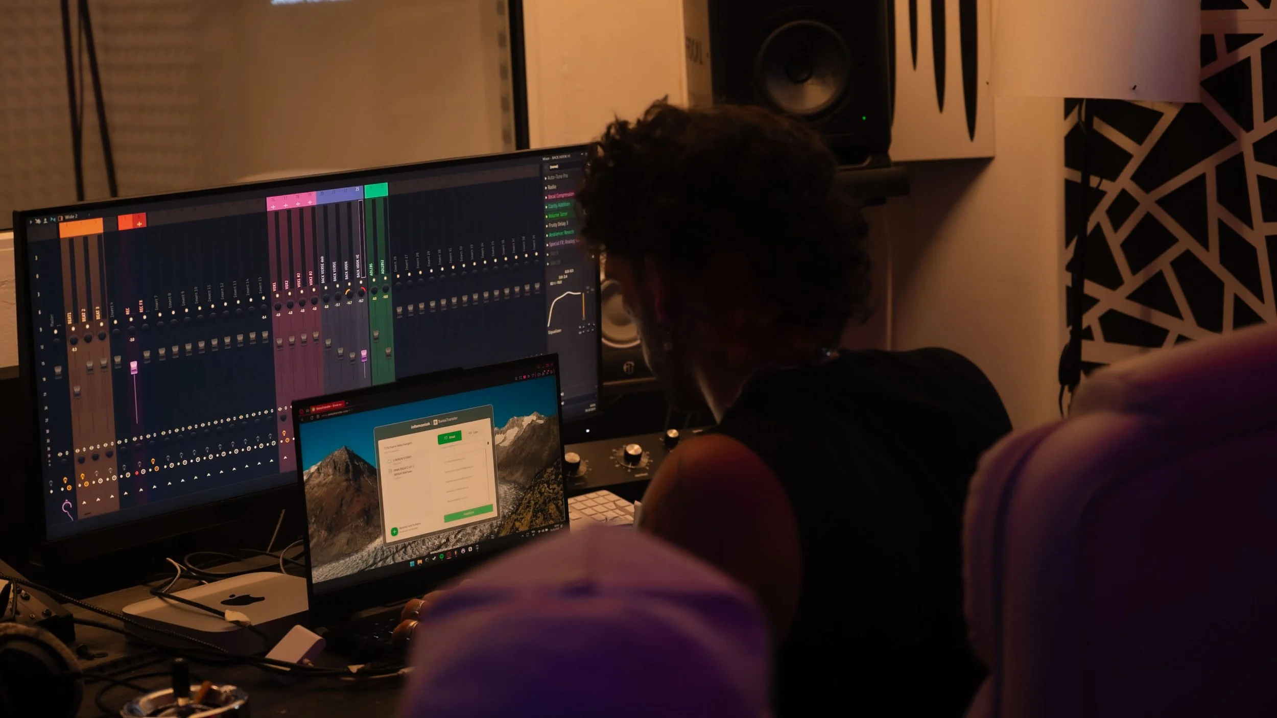 Un homme travaille sur un ordinateur avec un logiciel de production musicale. Il y a une grande enceinte et un écran supplémentaire en arrière-plan dans une pièce sombre.