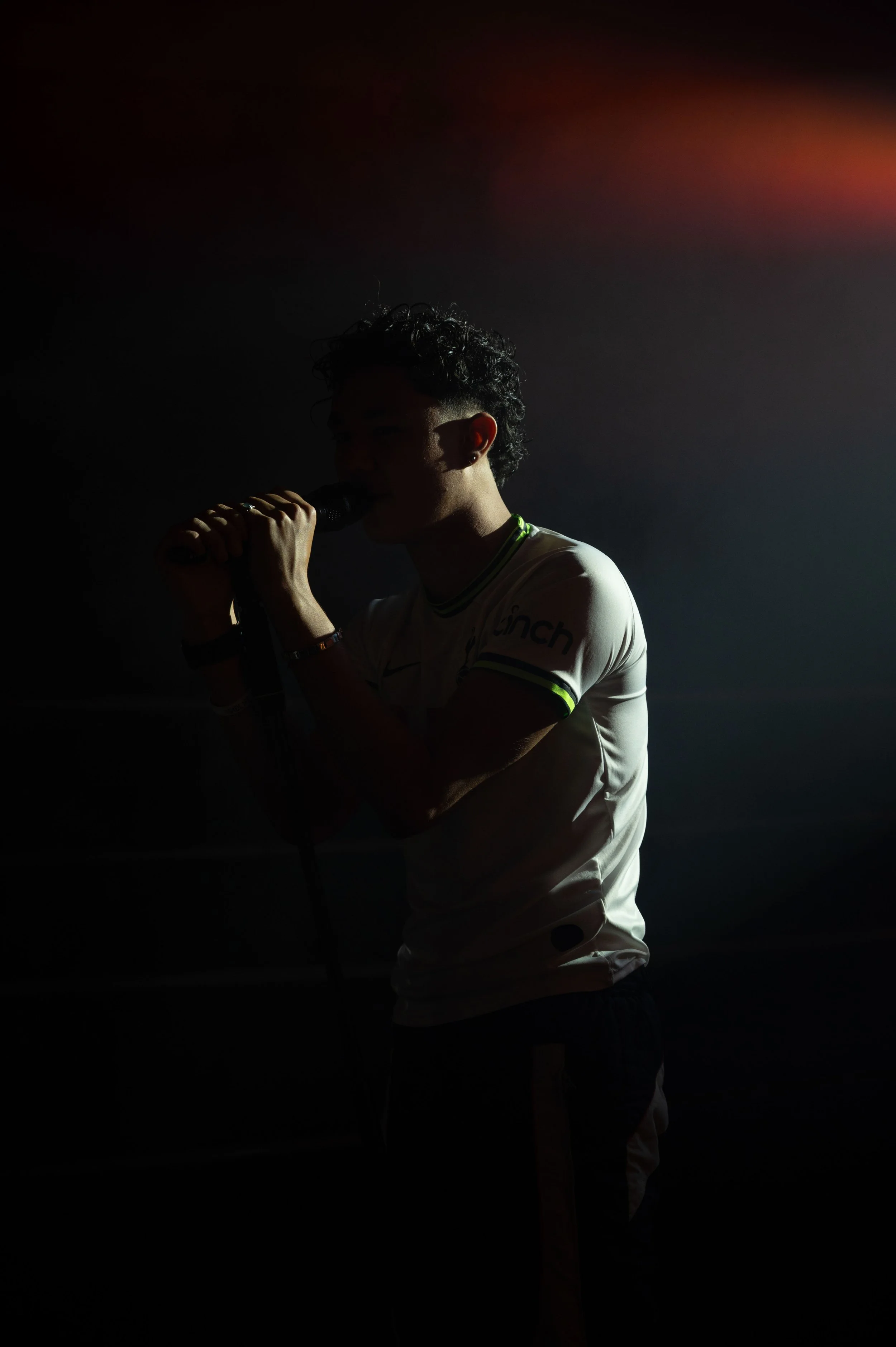 Une personne avec des cheveux bouclés, portant un t-shirt blanc et tenant un microphone, est en train de chanter ou parler dans une scène sombre avec un éclairage minimal.