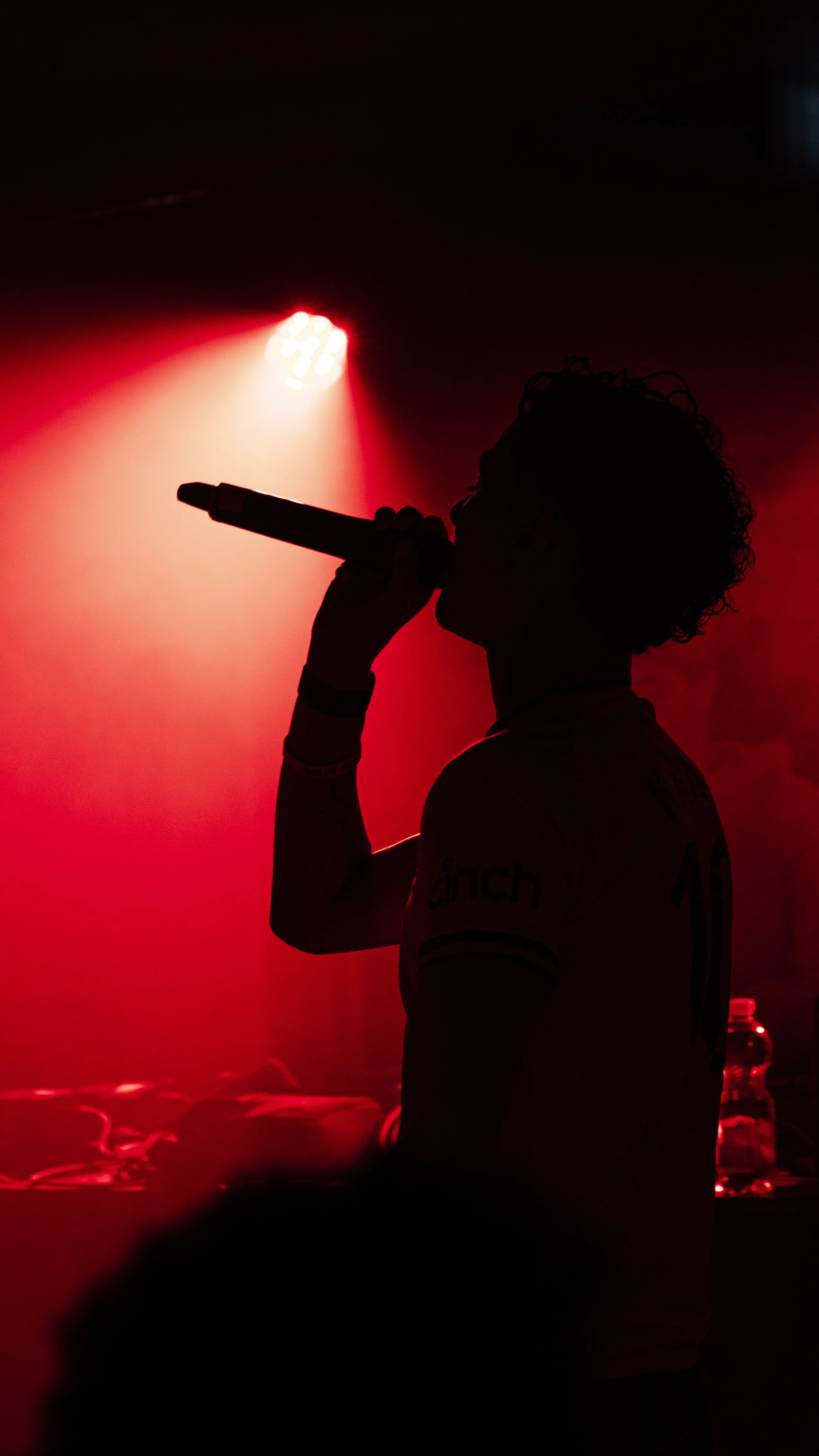 Silhouette d'une personne chantant ou parlant dans un microphone, éclairée par une lumière rouge dans une ambiance sombre.