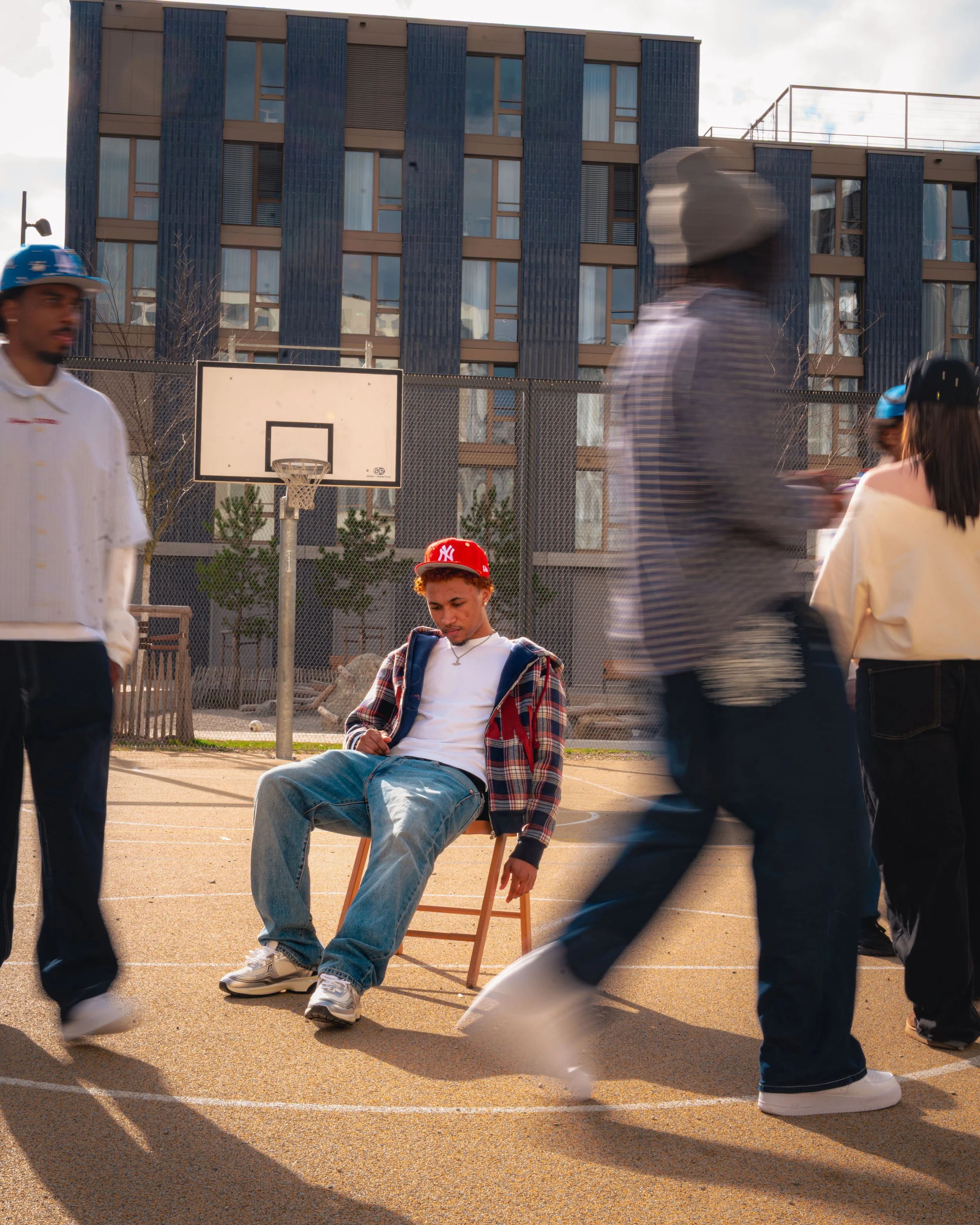 Une scène dans une cour de sport en plein air avec plusieurs jeunes. Au centre, un garçon assis sur une chaise regarde son téléphone, portant un hoodie à carreaux, un t-shirt blanc, des jeans et des baskets. Autour, plusieurs autres jeunes marchent o