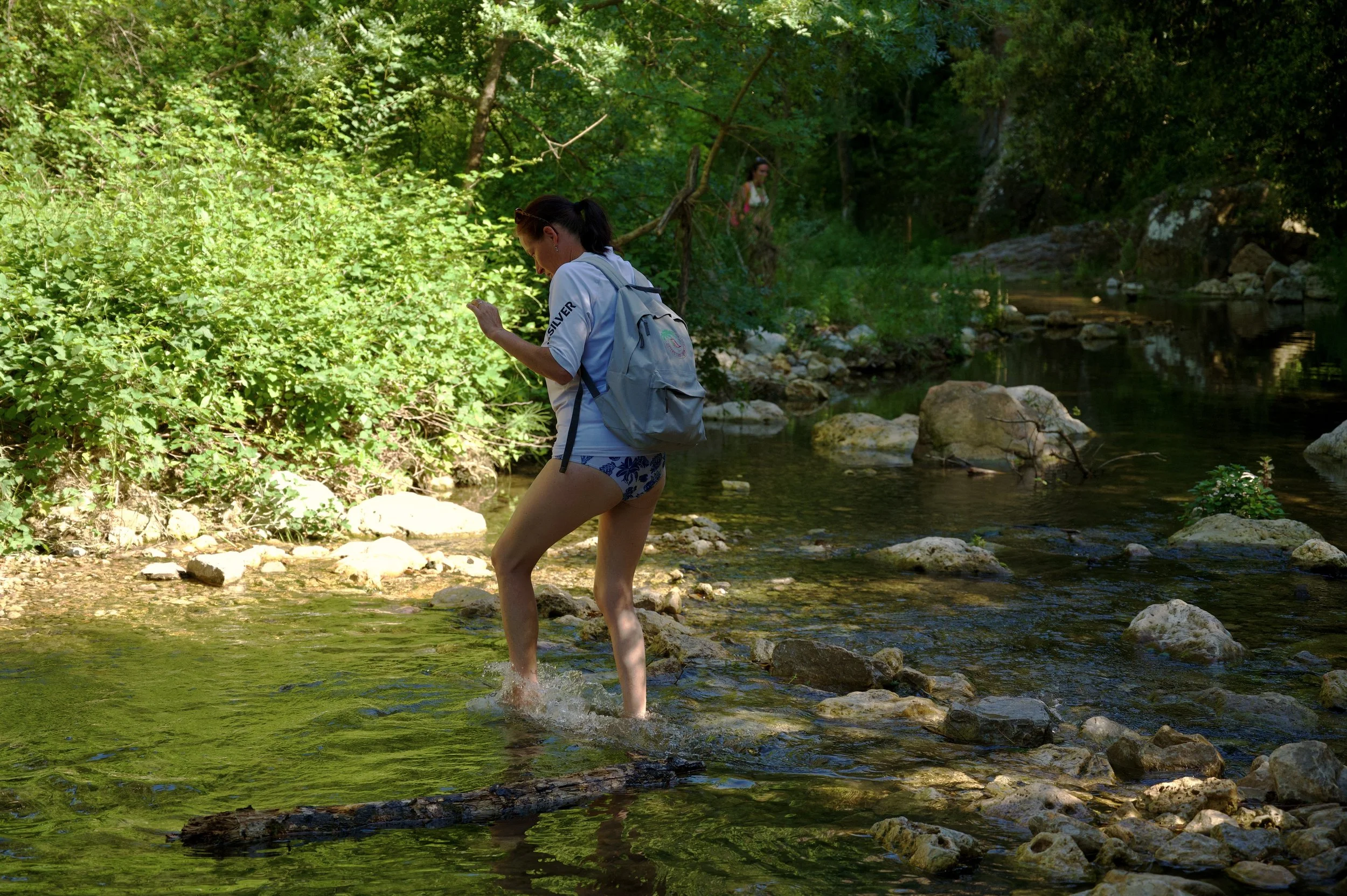 Femme traversant un ruisseau peu profond en été, portant un sac à dos et des vêtements décontractés, entourée de végétation verte dense.