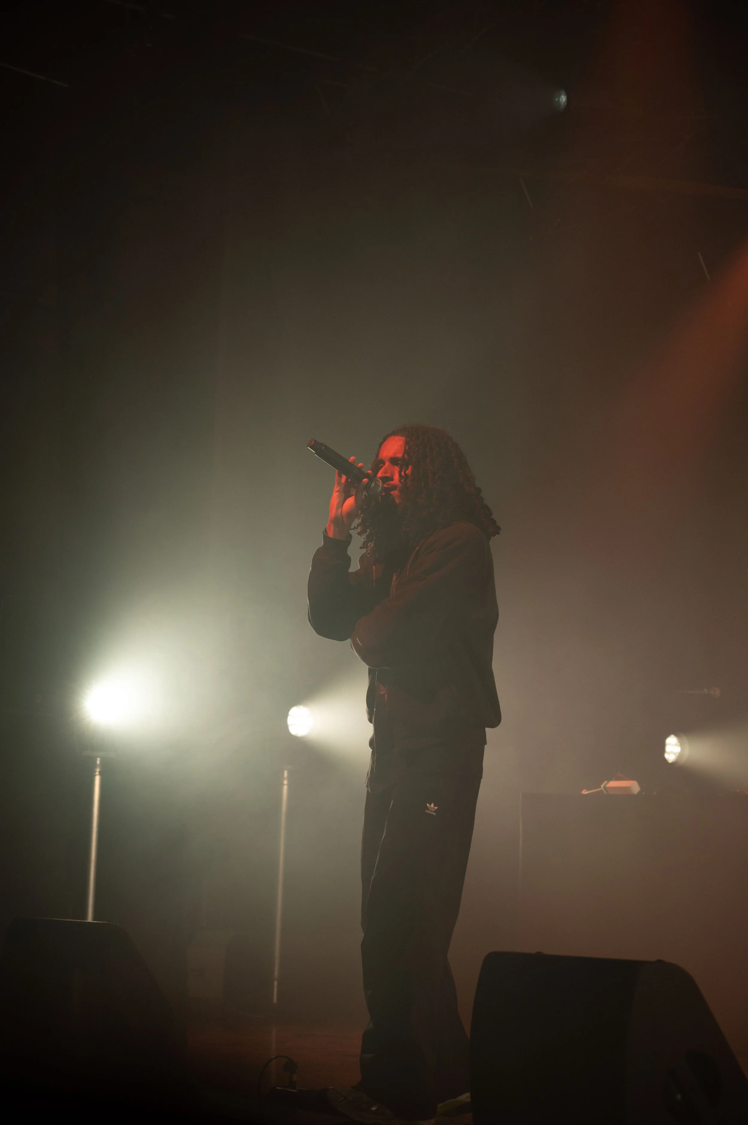 Un homme rasé de près avec des cheveux bouclés, portant une veste noire et un pantalon noir, chante en micro dans une scène de concert avec des lumières tamisées et de la fumée, arrière-plan sombre.