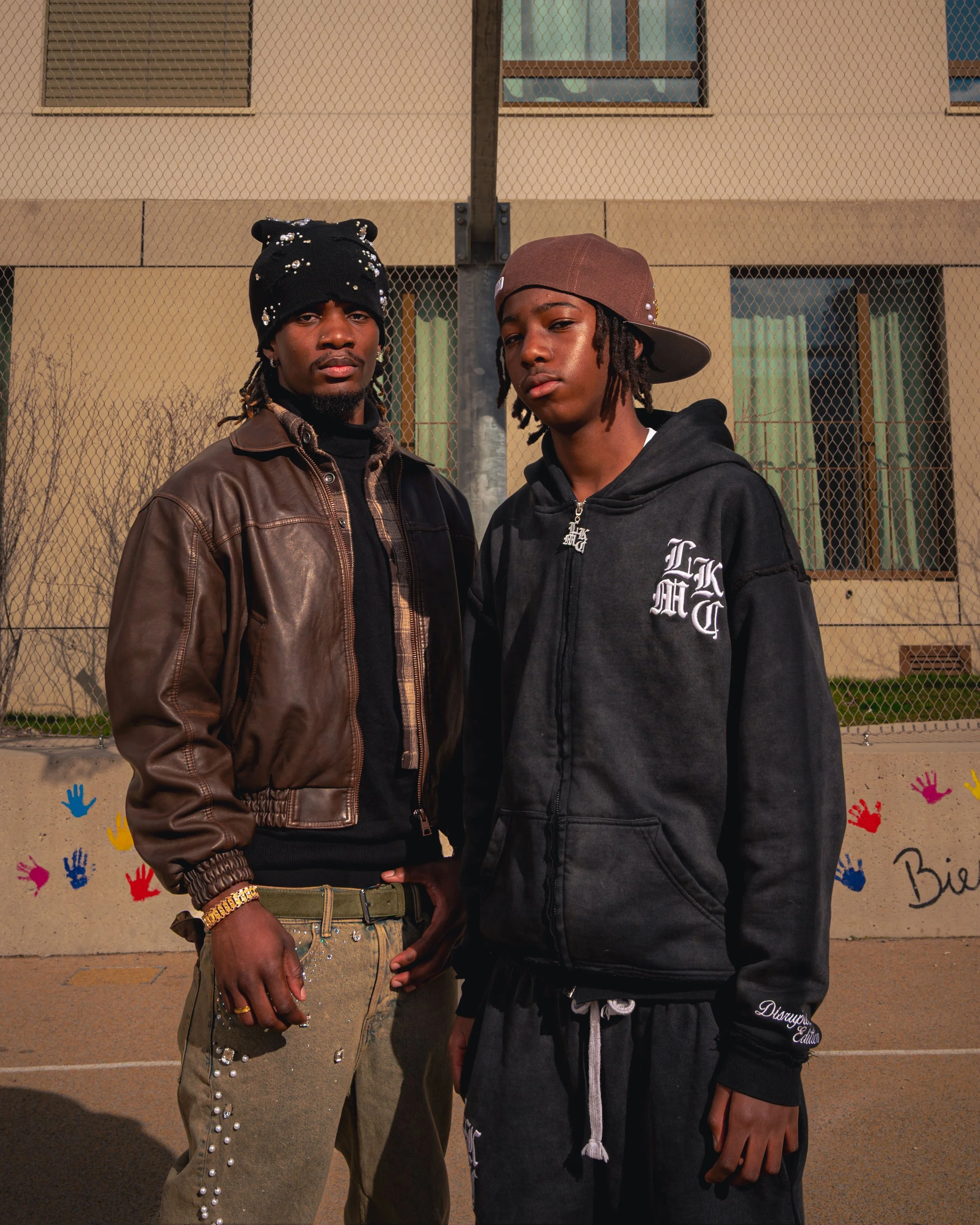Deux jeunes hommes posent devant une clôture de terrain de basket-ball avec un bâtiment en arrière-plan, un en costume chic avec une casquette et un autre en vêtements décontractés, tous deux regardant vers l'objectif.