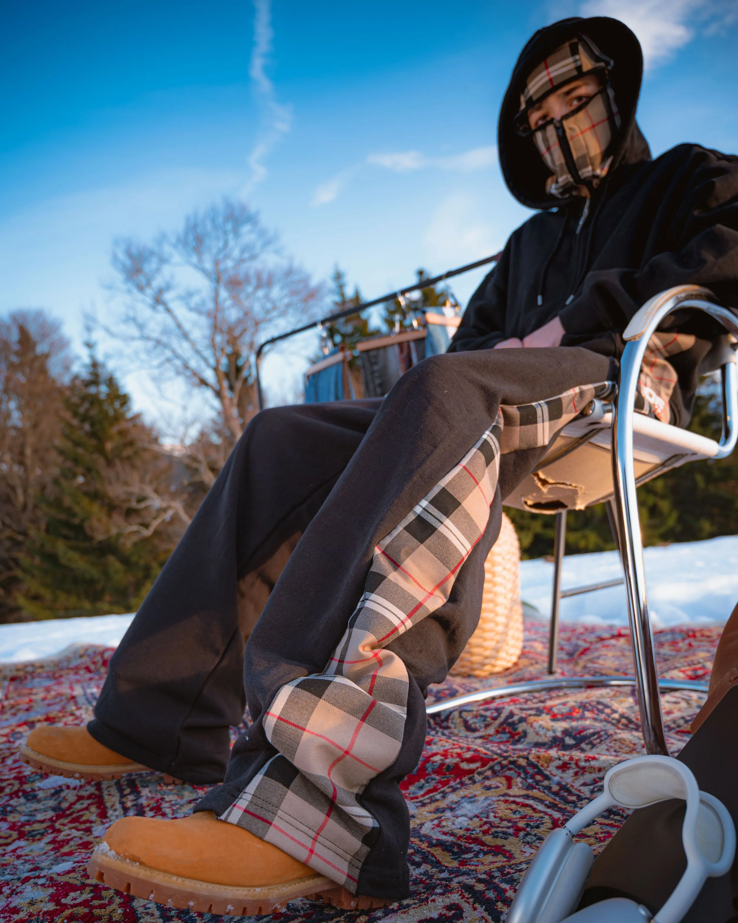 Personne dans un fauteuil roulant portant une capuche et un masque, dans un extérieur avec un ciel bleu et des arbres dénudés, assise sur un tapis persan, portant des bottes en cuir beige.