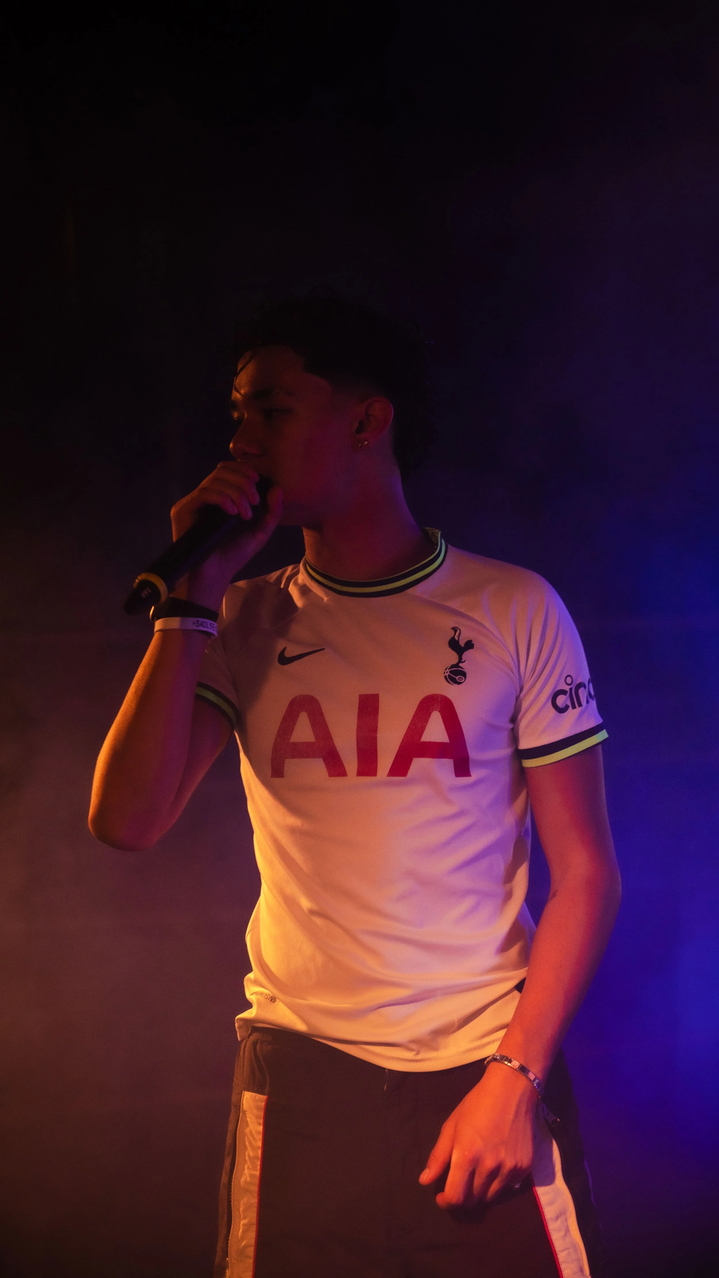 Un jeune homme en maillot de football blanc avec le logo des Spurs et le sponsor AIA, tenant un micro, dans une ambiance sombre avec un éclairage violet et chaud.