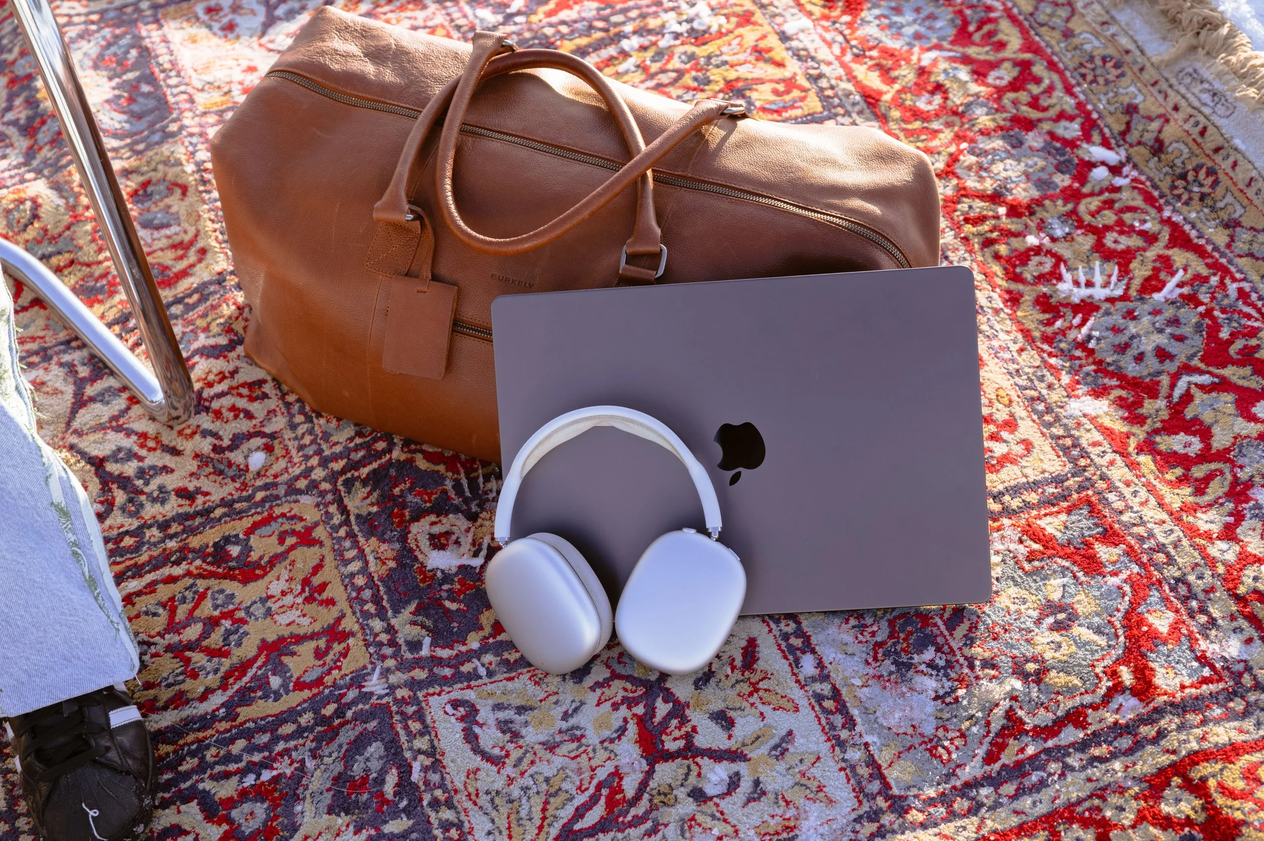 Sac de voyage en cuir marron sur un tapis persan, avec un ordinateur portable, des écouteurs et une paire de chaussures blanches autour.