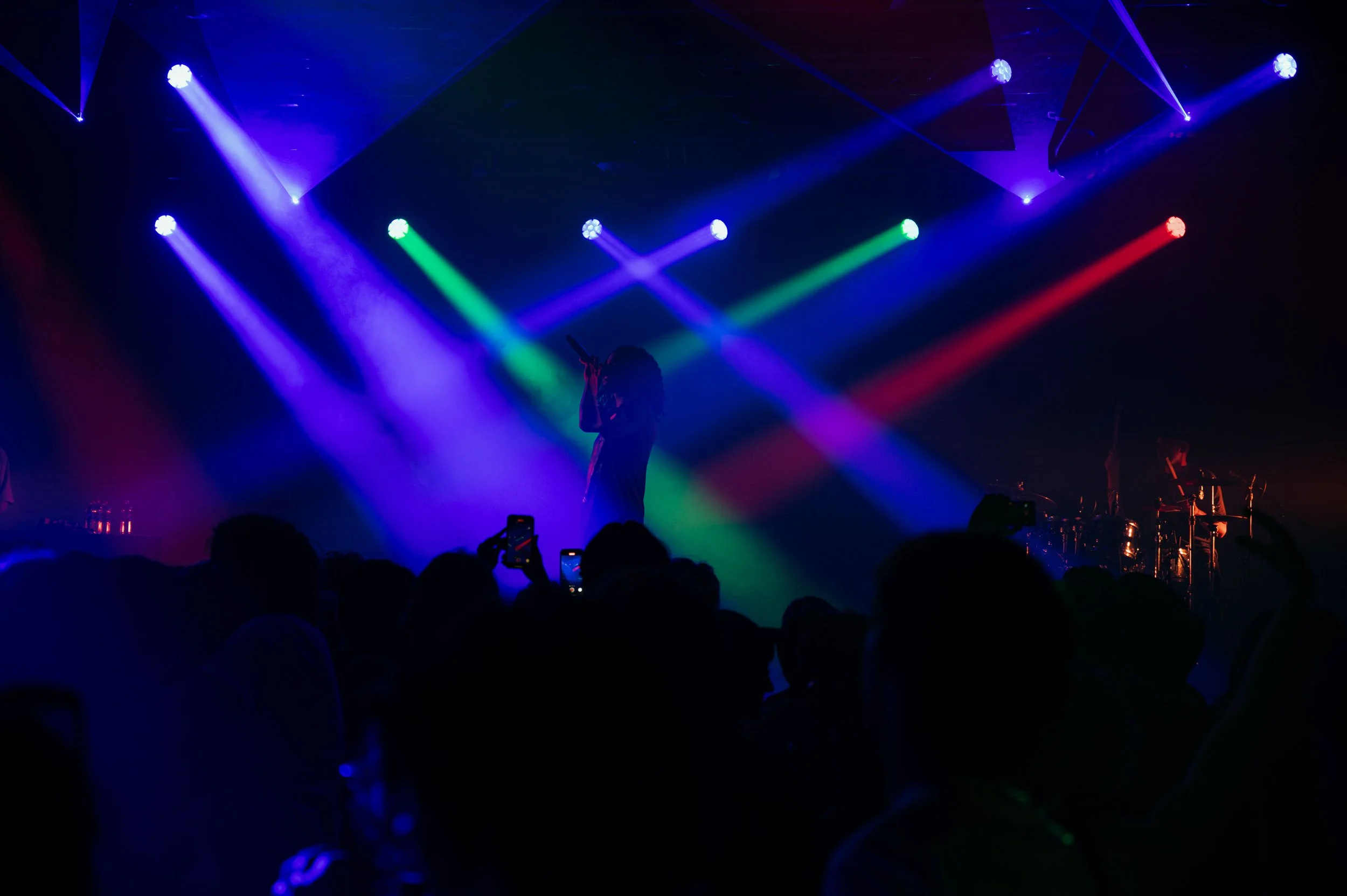 Une scène de concert avec un éclairage coloré de lumières violettes, vertes et rouges, et une silhouette de personne chantant ou parlant au microphone, entourée par une foule vue en silhouette