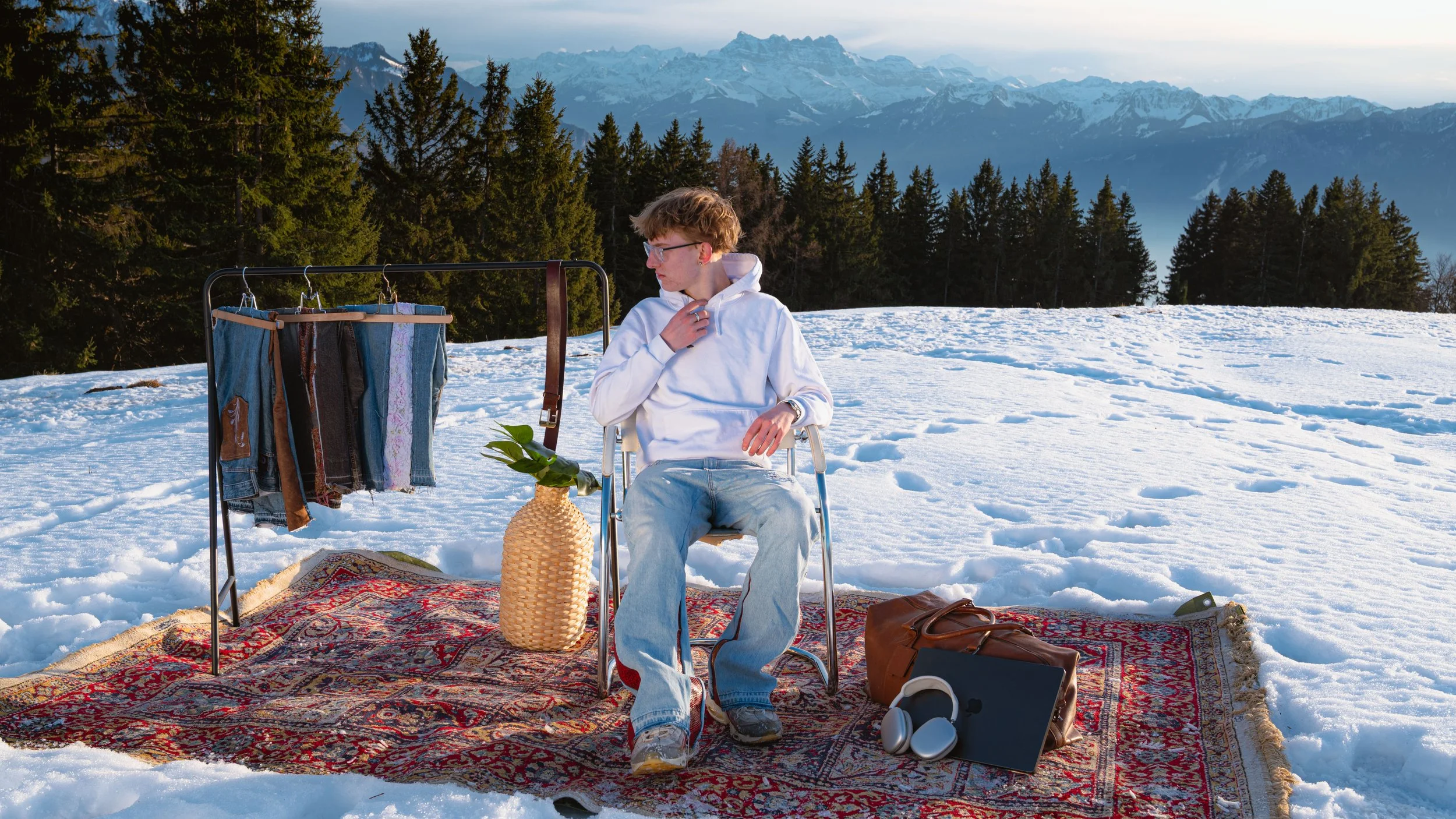 Jeune homme assis dans un fauteuil dans la neige, avec une étagère à vêtements, un tapis, une plante et des écouteurs, devant une forêt et des montagnes enneigées.