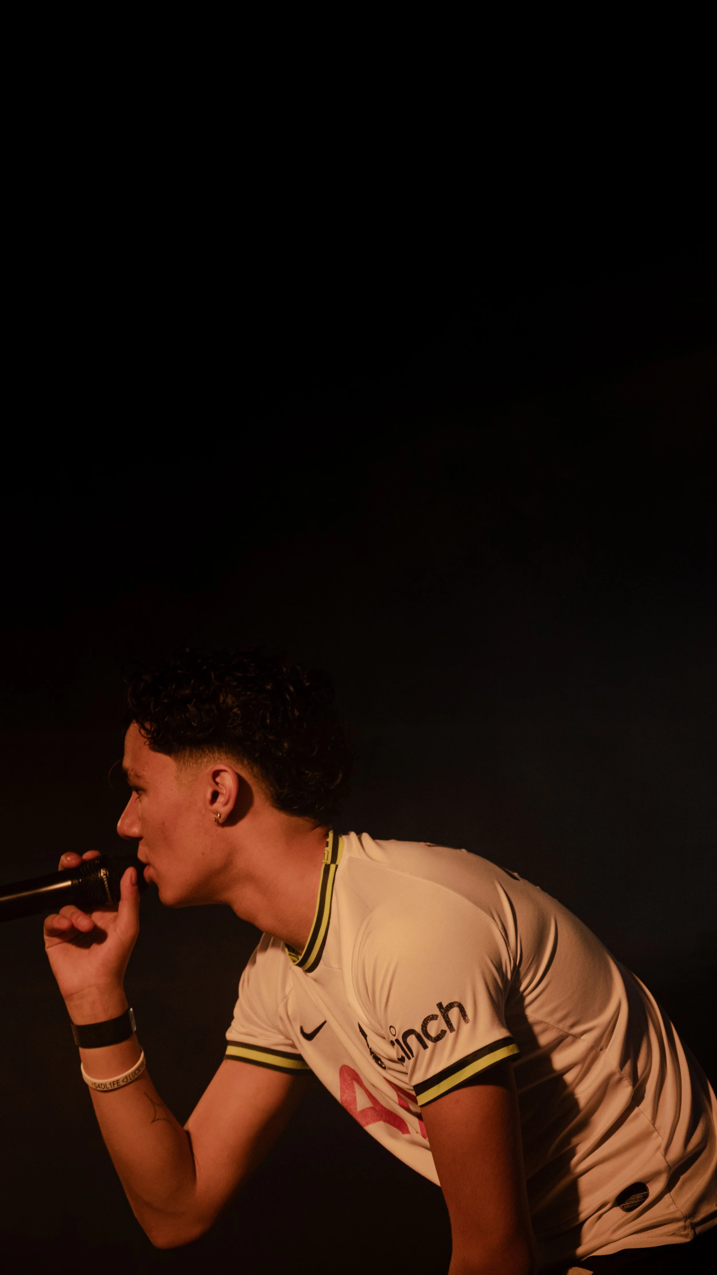 Jeune homme avec des cheveux bouclés courts, portant un maillot de football blanc avec des détails noirs et jaunes, tenant un microphone près de la bouche dans un décor sombre.
