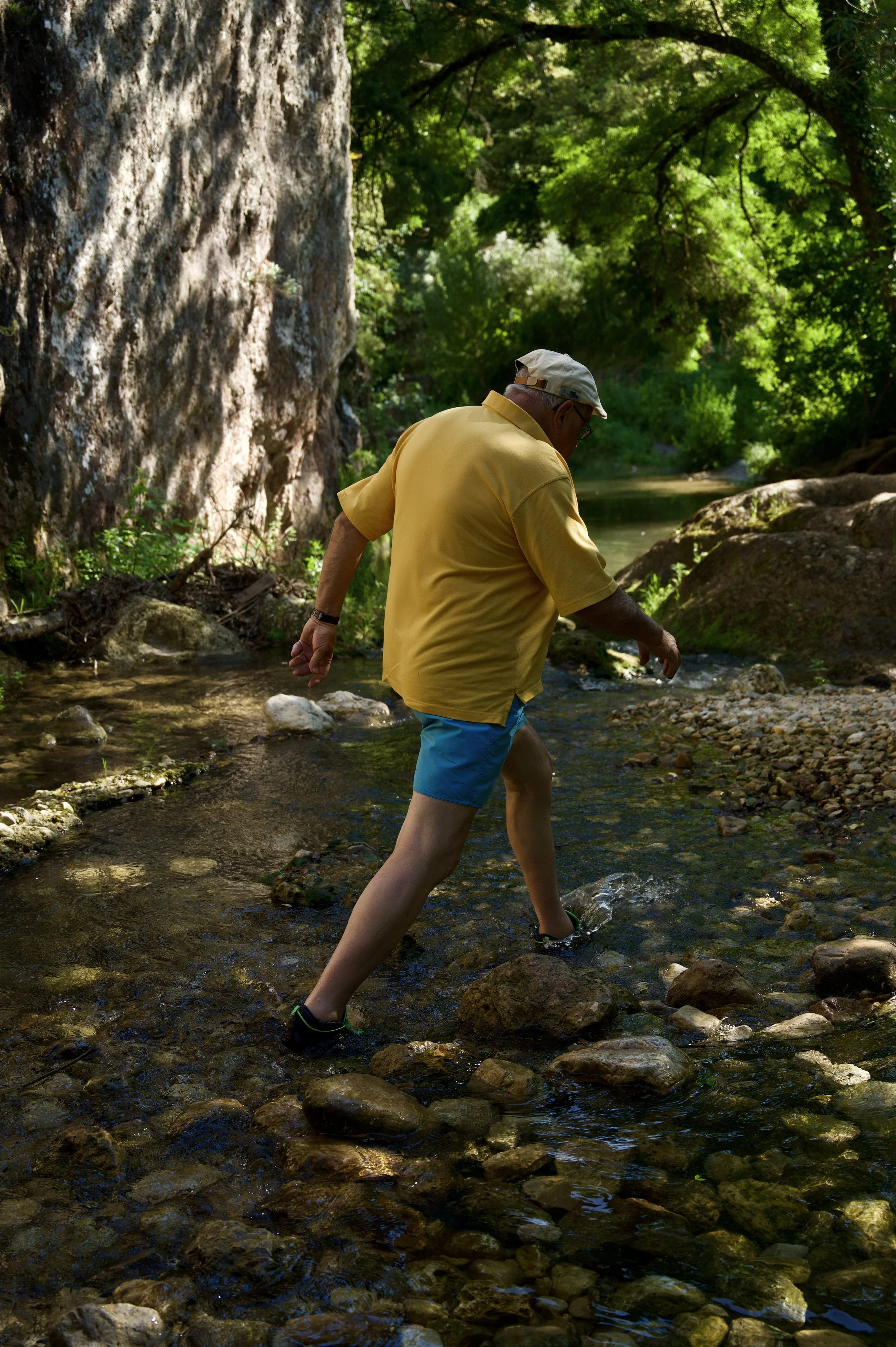 Un homme âgé portant un chapeau blanc, un t-shirt jaune et un short bleu, traversant une rivière dans une forêt dense verte, en marchant sur des rochers et en touchant l'eau.