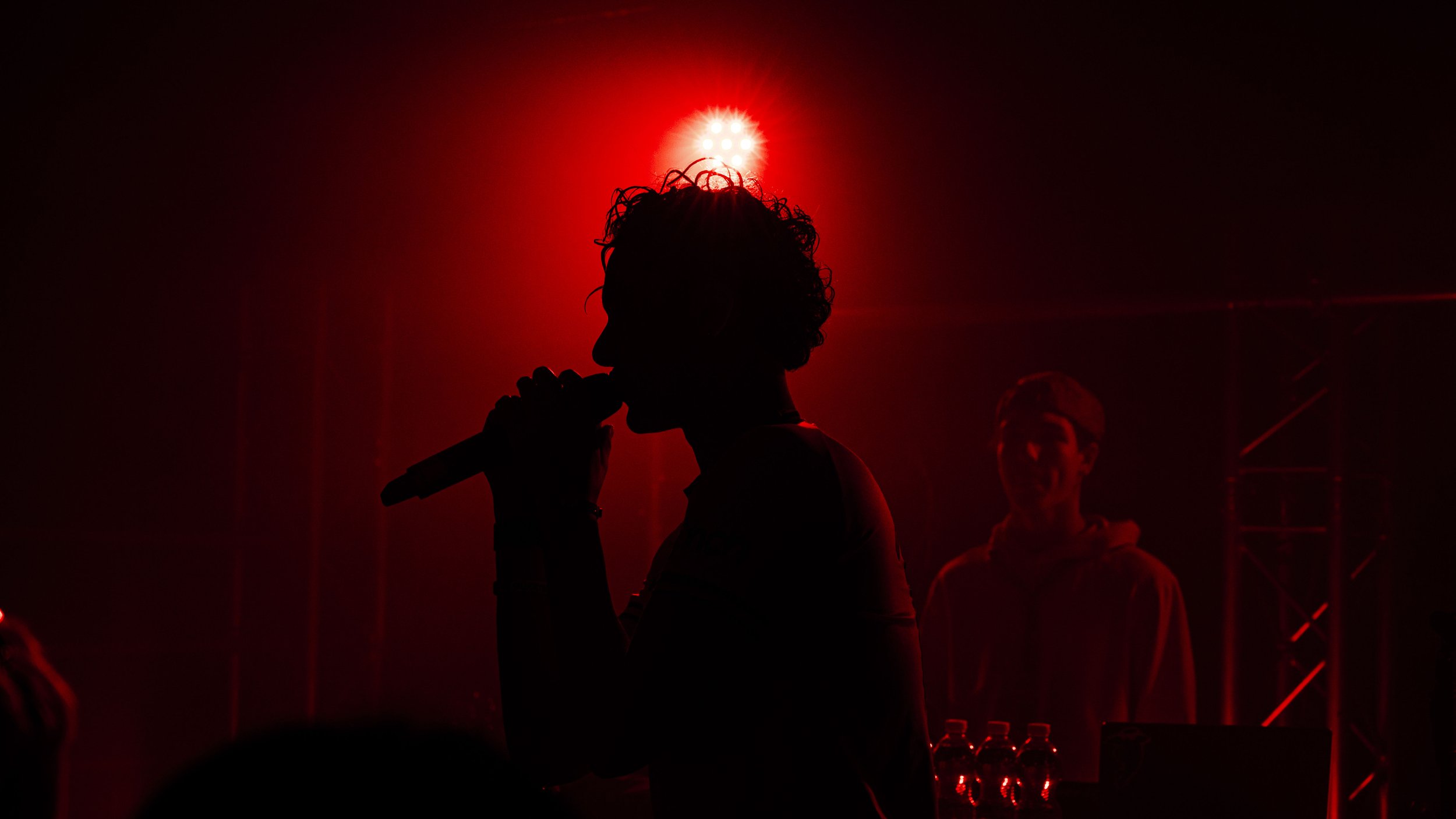Silhouette d'une personne chantant ou parlant dans un microphone sous un éclairage rouge intense. Seconde personne en arrière-plan souriant, dans une ambiance de concert ou de scène.