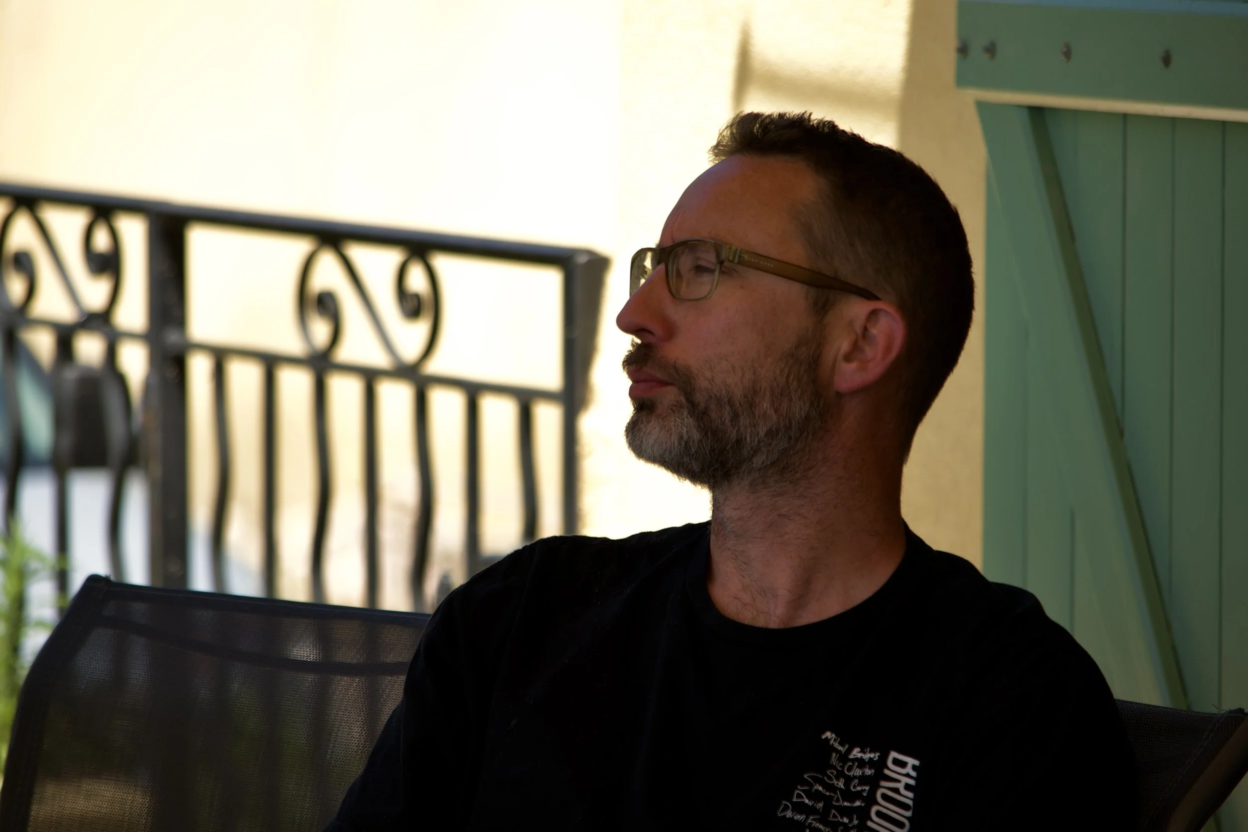 Homme avec lunettes, barbe, assis en plein air, regardant vers la droite, derrière un balcon en métal noir et un mur vert à l'arrière-plan.