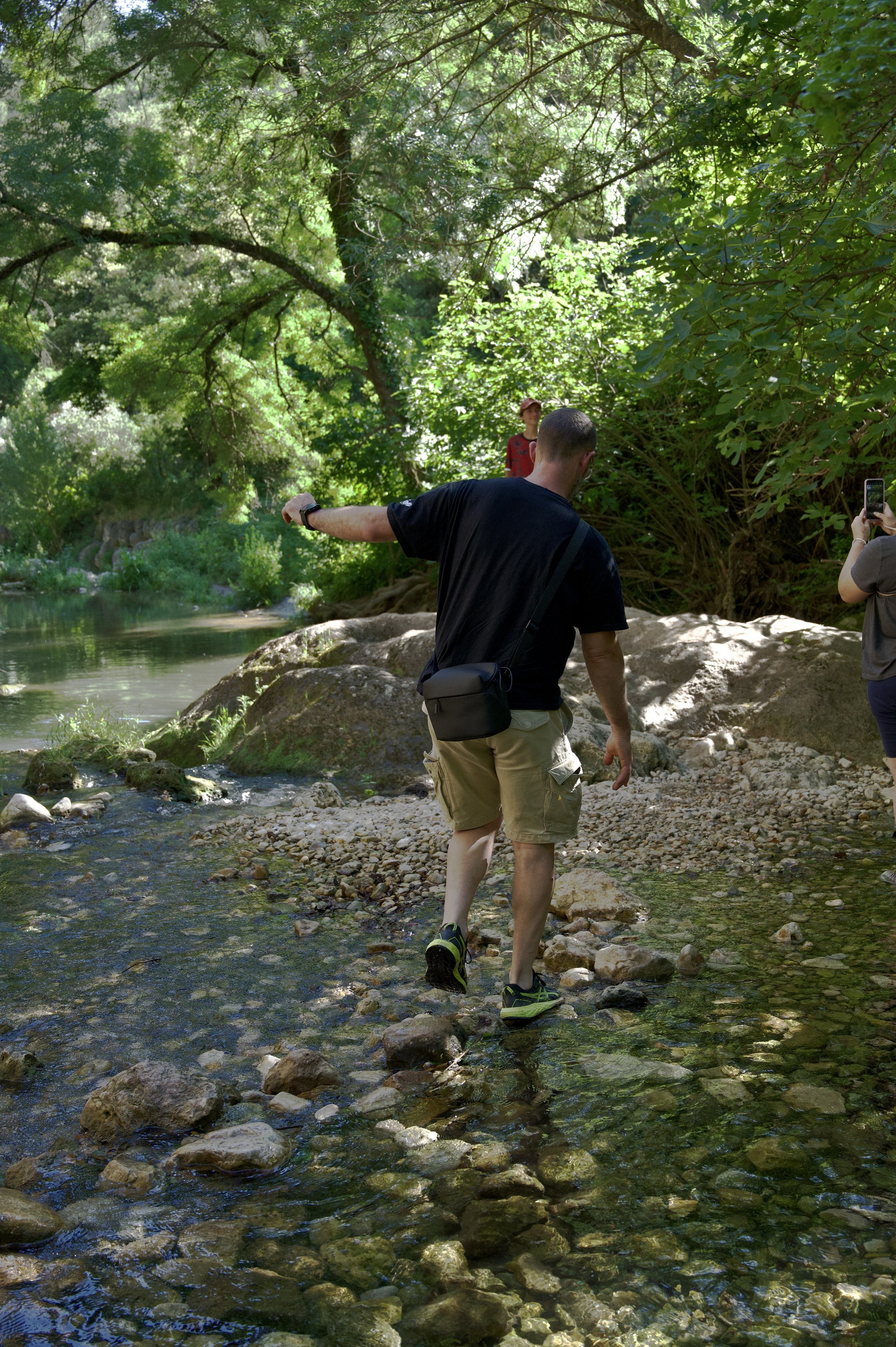 Homme traversant une rivière peu profonde avec des cailloux, entouré de végétation dense et de grands arbres dans un environnement naturel verdoyant.