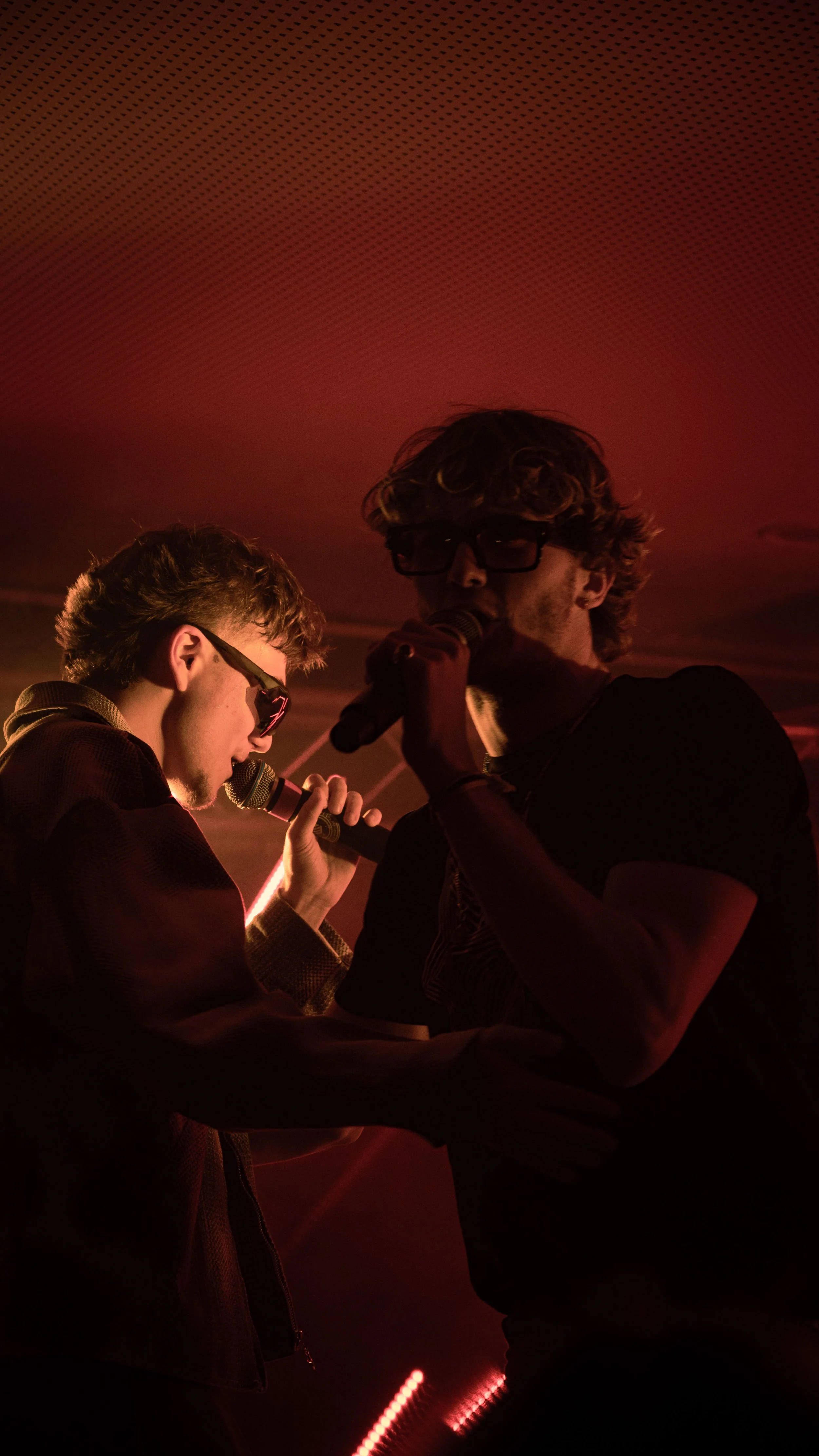 Deux jeunes hommes chantent avec des microphones dans un environnement sombre éclairé par une lumière rouge, l'un porte des lunettes et l'autre des lunettes de soleil.