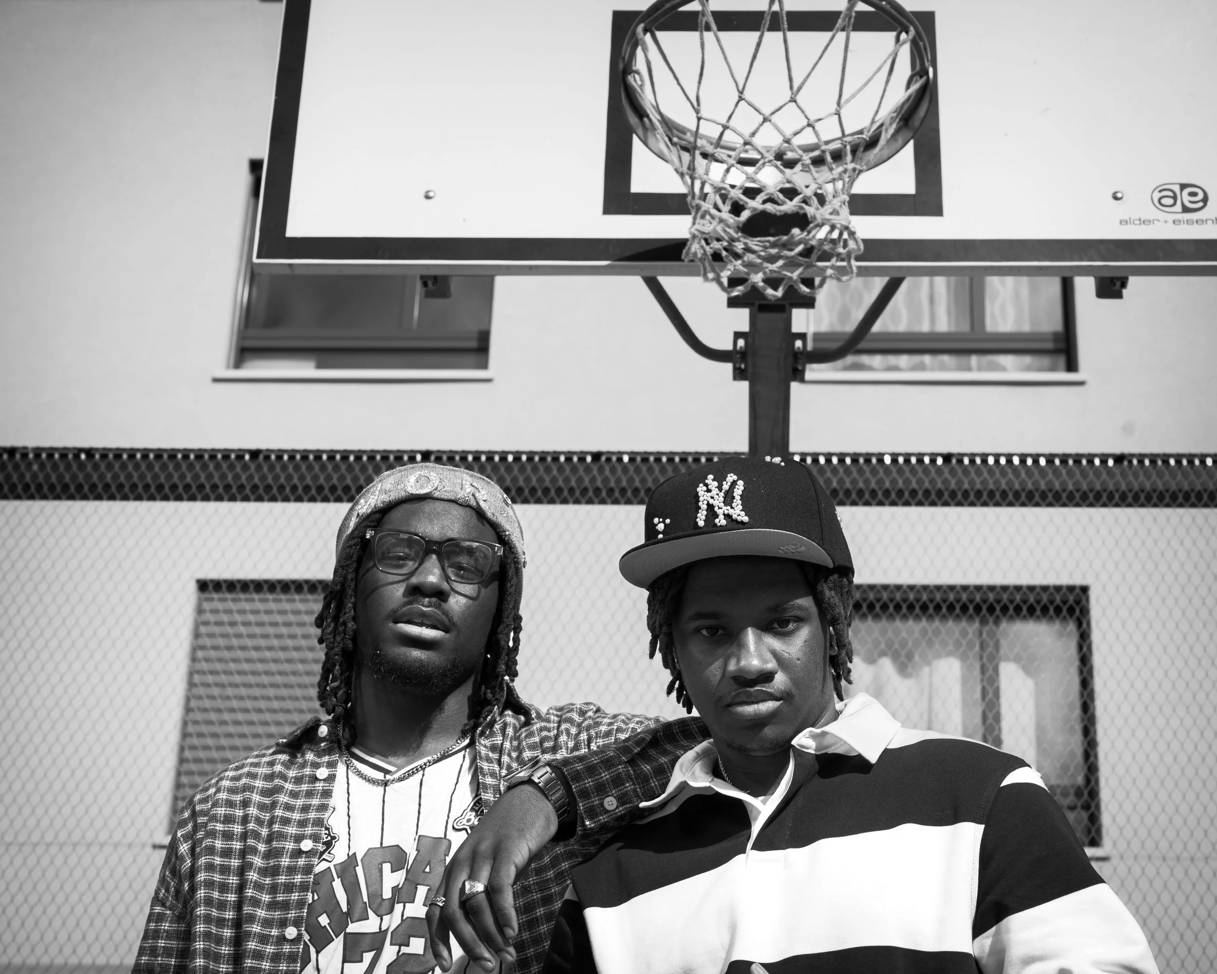 Deux jeunes hommes posent devant un panier de basketball suspendu. L'un porte une casquette noire avec les lettres 'NY' et une chemise à carreaux, l'autre porte une chemise à rayures. La photo est en noir et blanc.