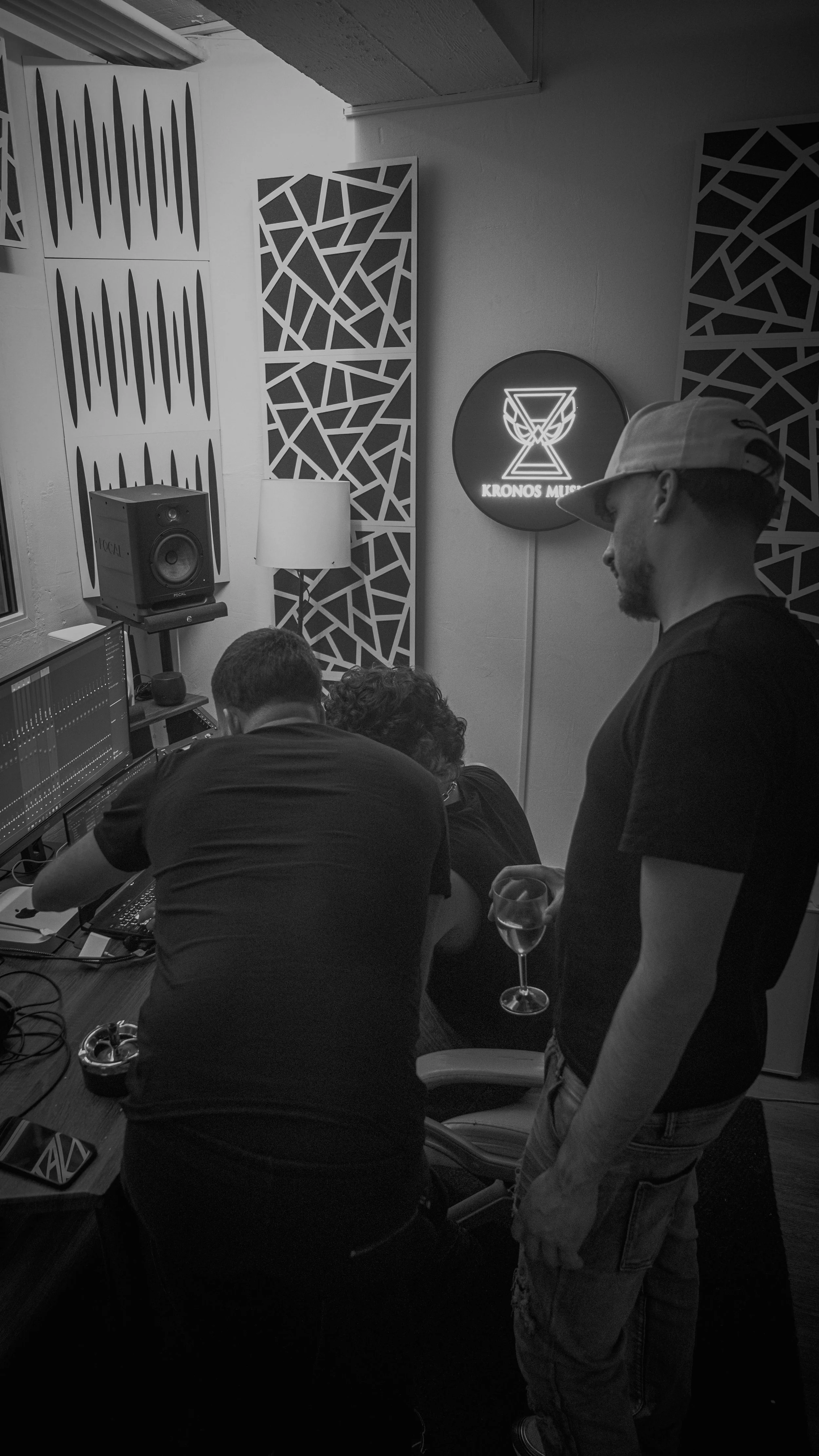 Une photo en noir et blanc montrant un groupe de personnes dans un studio de production musicale. Deux personnes semblent concentrées devant un ordinateur. Une personne debout à droite, portant une casquette et un t-shirt noir, tient un verre de vin 