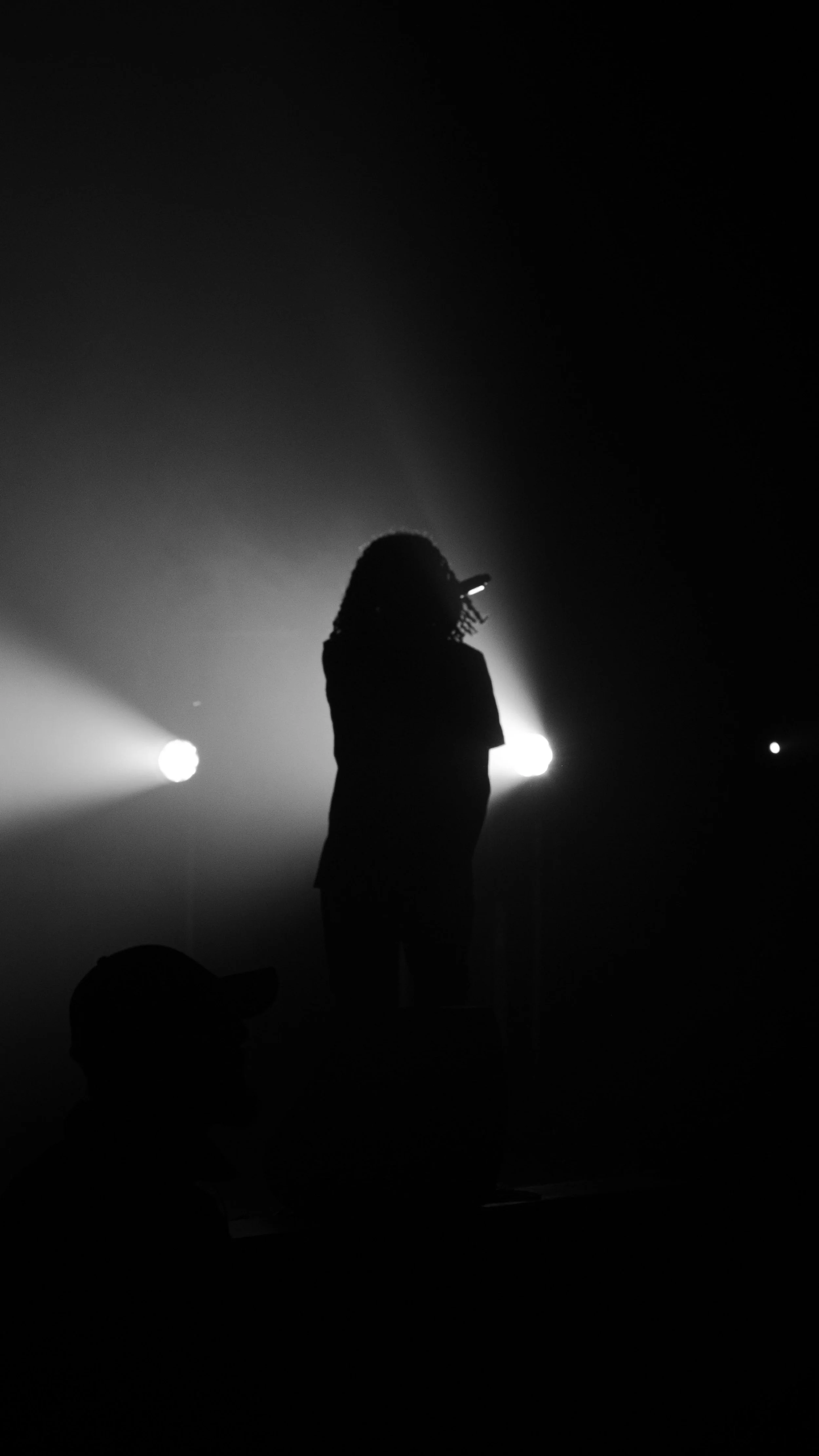 Silhouette d'une personne avec un micro, éclairée par des lumières à l'arrière-plan, en noir et blanc.
