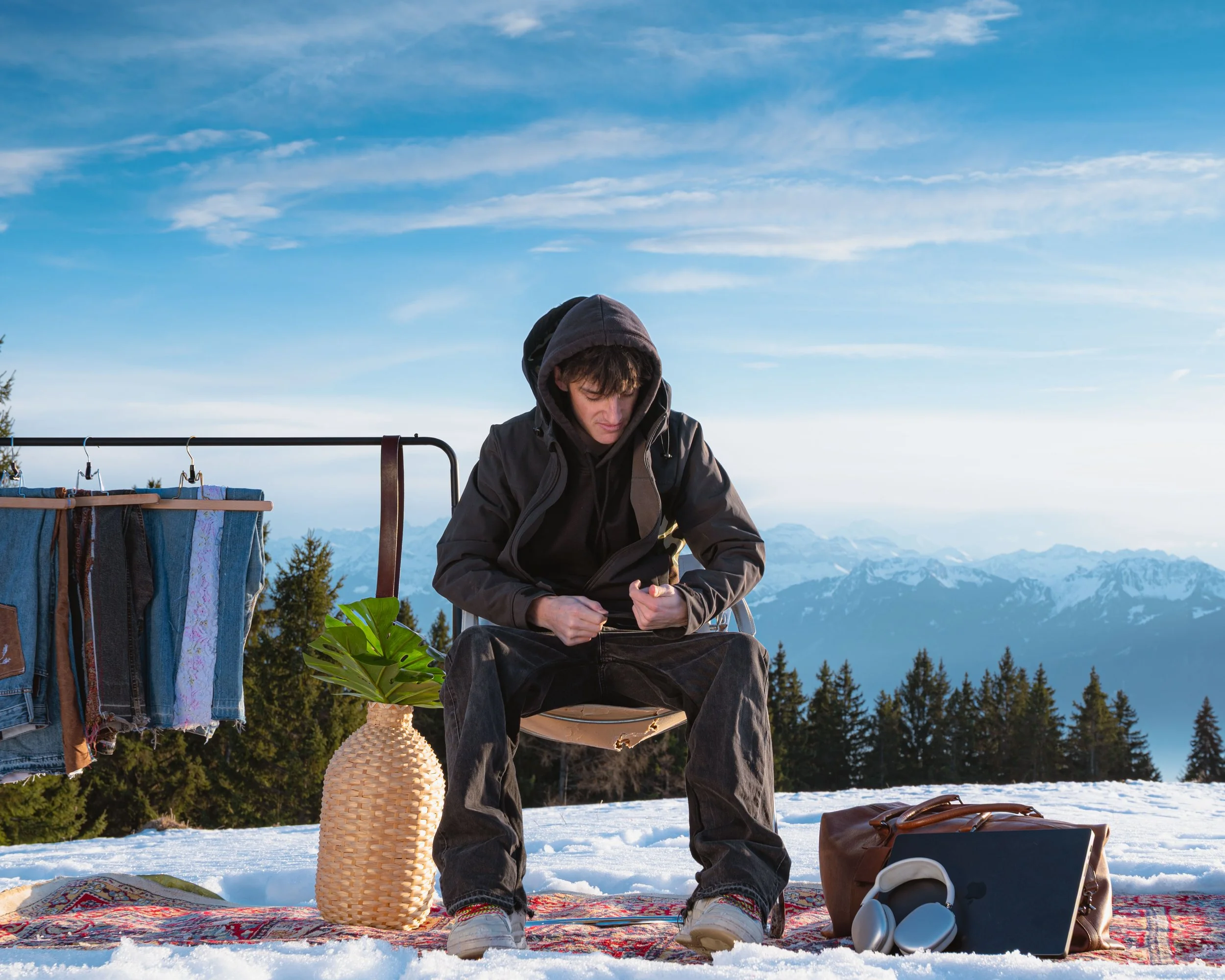 Jeune homme assis dans un paysage enneigé, portant une veste à capuche, avec des montagnes et un ciel bleu en arrière-plan, entouré d'objets de camping et de vêtements suspendus, au soleil.