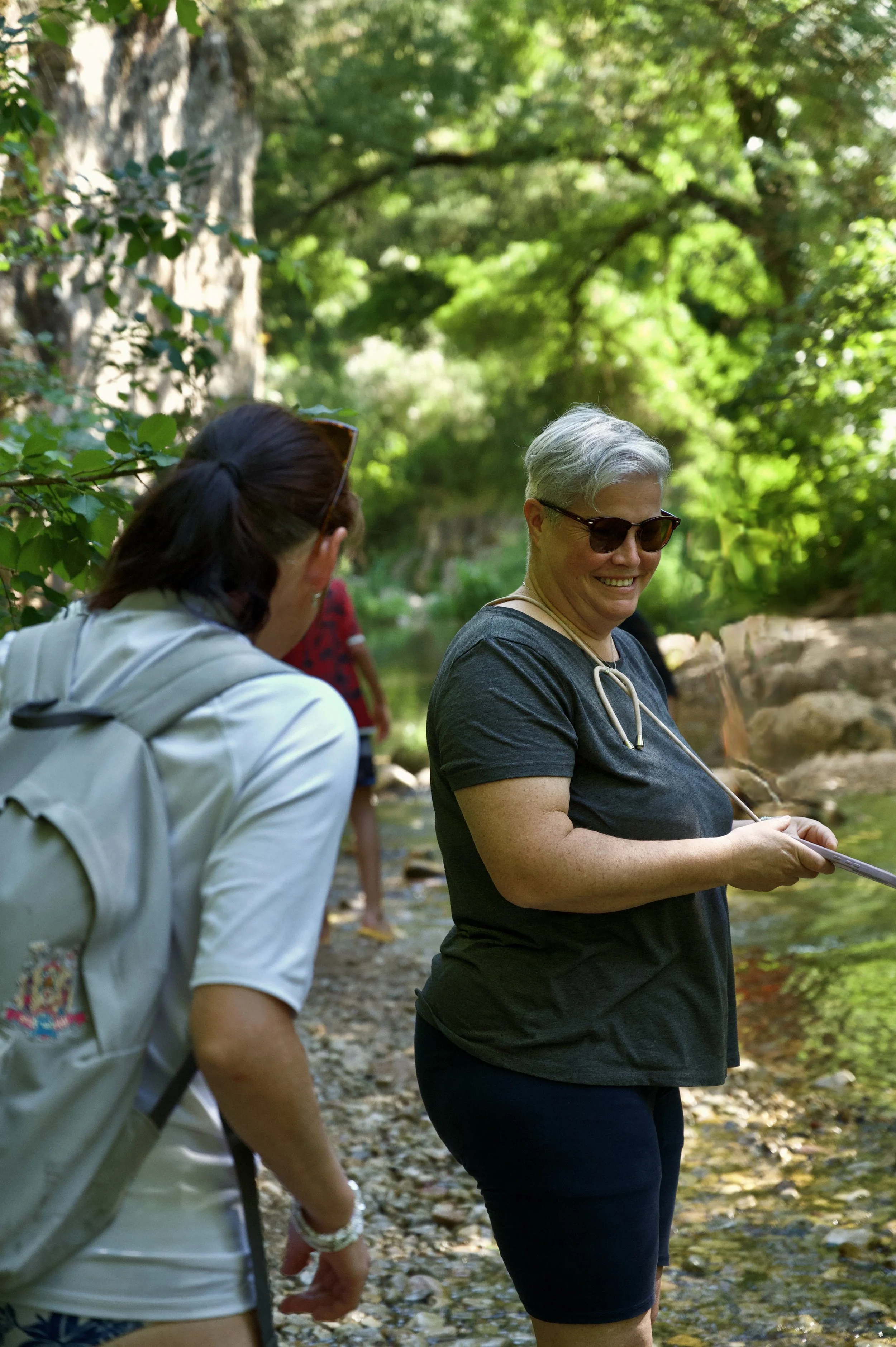 Deux femmes debout près d'une rivière dans une forêt verte, discutant et souriant, l'une porte des lunettes de soleil et l'autre un sac à dos.